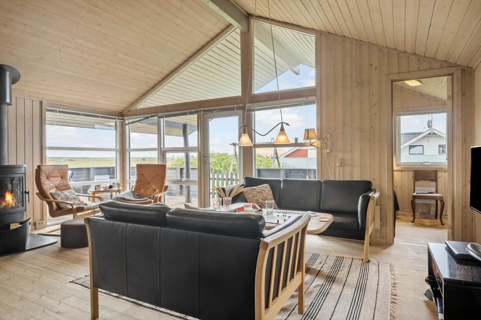 Wohnzimmer mit Holzdecke, Sofa, Sessel und Kamin. Große Fenster bieten Blick auf die Landschaft.