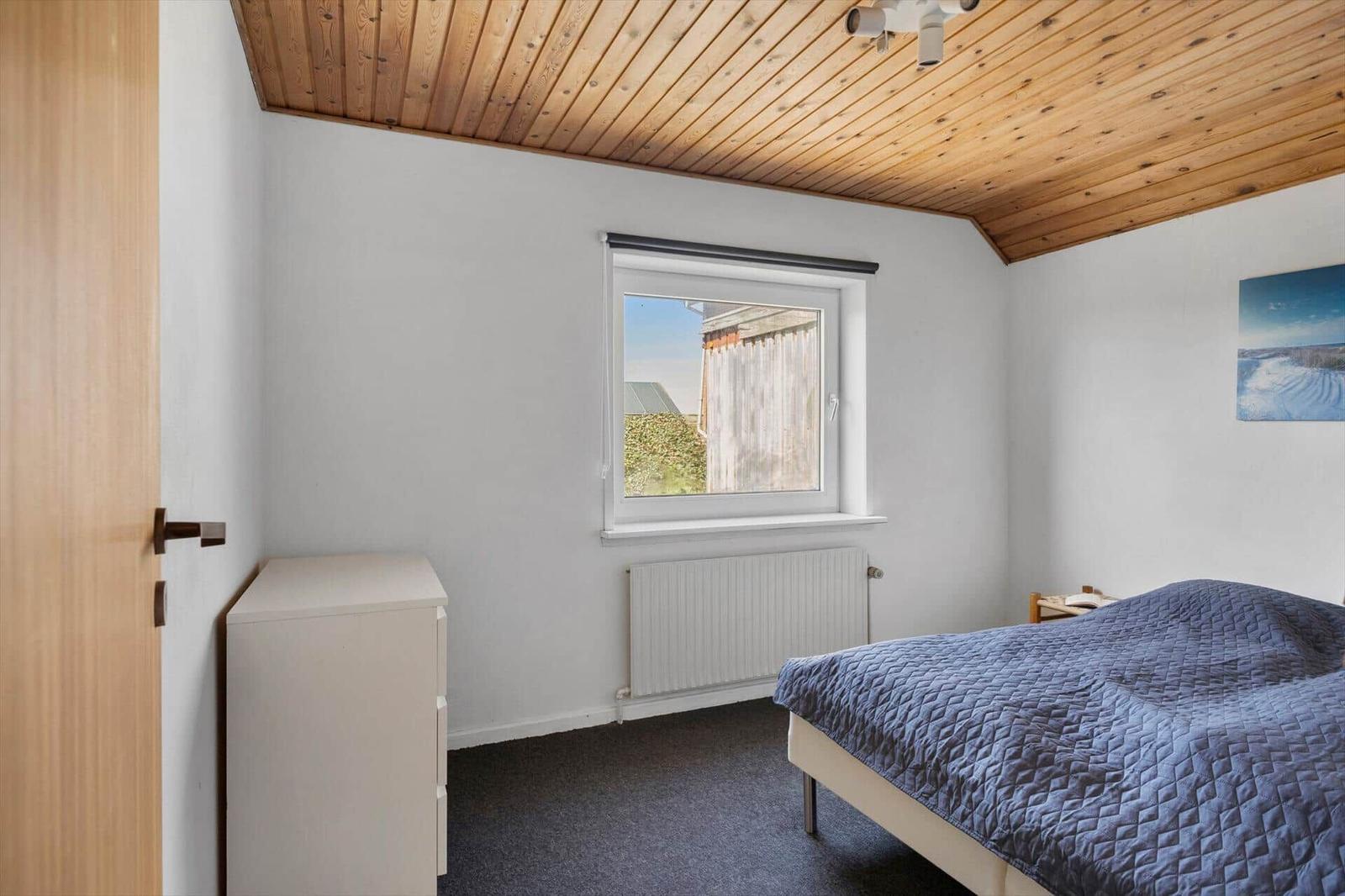 Schlafzimmer mit Bett, Schrank und Fenster. Holzdecke und Wandheizung.