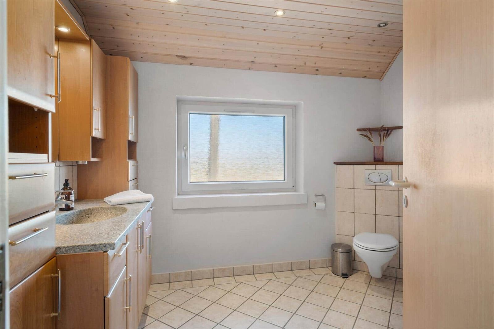 Badezimmer mit Holzdecke, Waschbecken und Toilette. Fenster mit Blick auf Wasser.