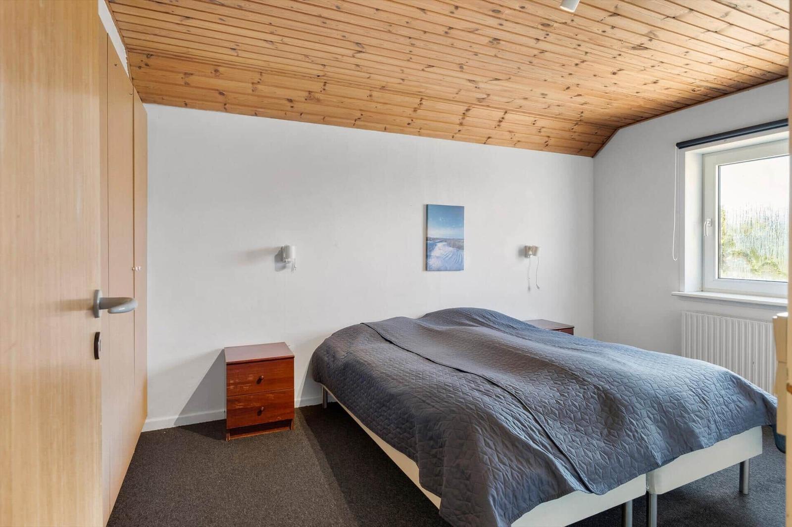 Schlafzimmer mit Bett, Nachttisch und Fenster. Holzdecke und Kleiderschrank sichtbar.
