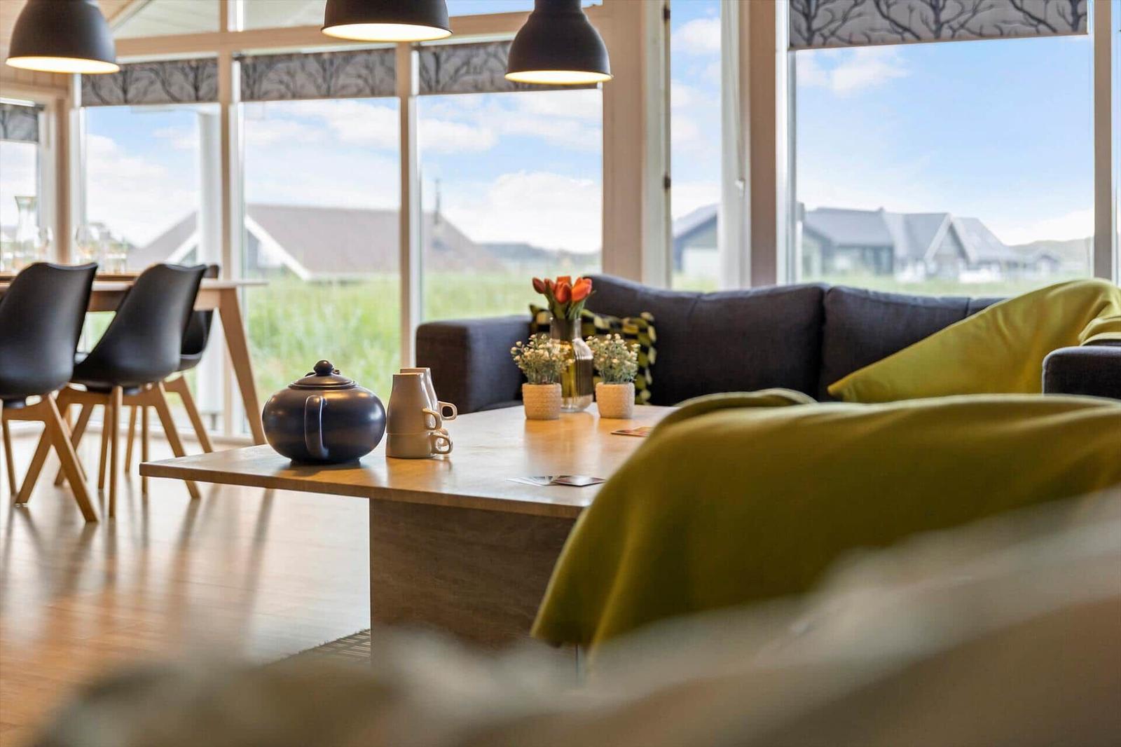 Ein Wohnzimmer mit Sofa, Tisch und großen Fenstern. Draußen ist eine grüne Landschaft zu sehen.