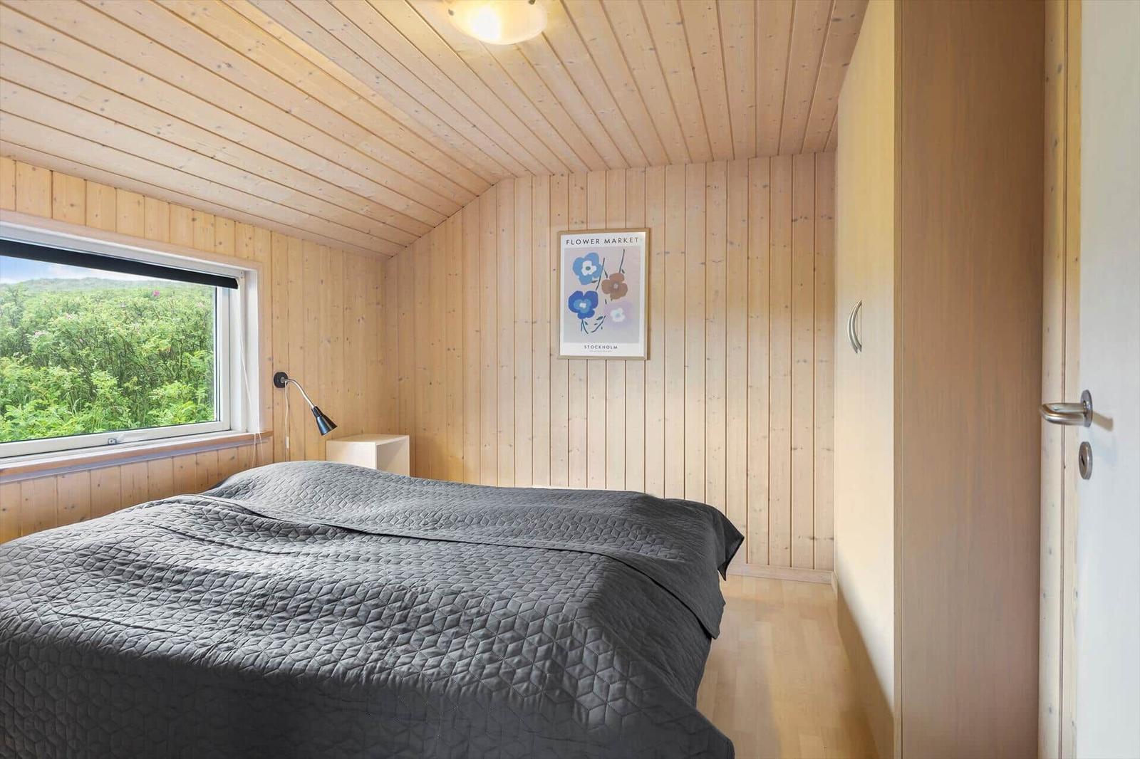 Schlafzimmer mit Holzwand und -decke, Bett mit grauer Bettdecke, Fenster mit Blick auf Grünflächen.