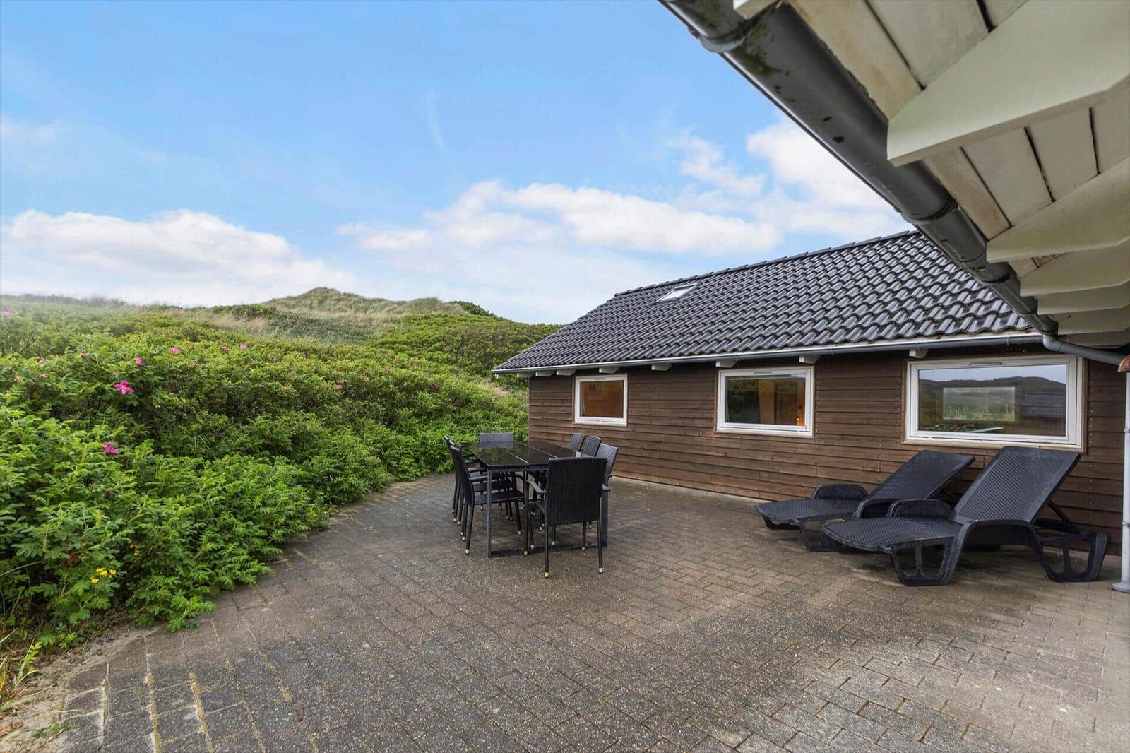 Terrasse mit Tisch, Stühlen und Liegestühlen neben einem Holzhaus mit Dach und Fenstern.