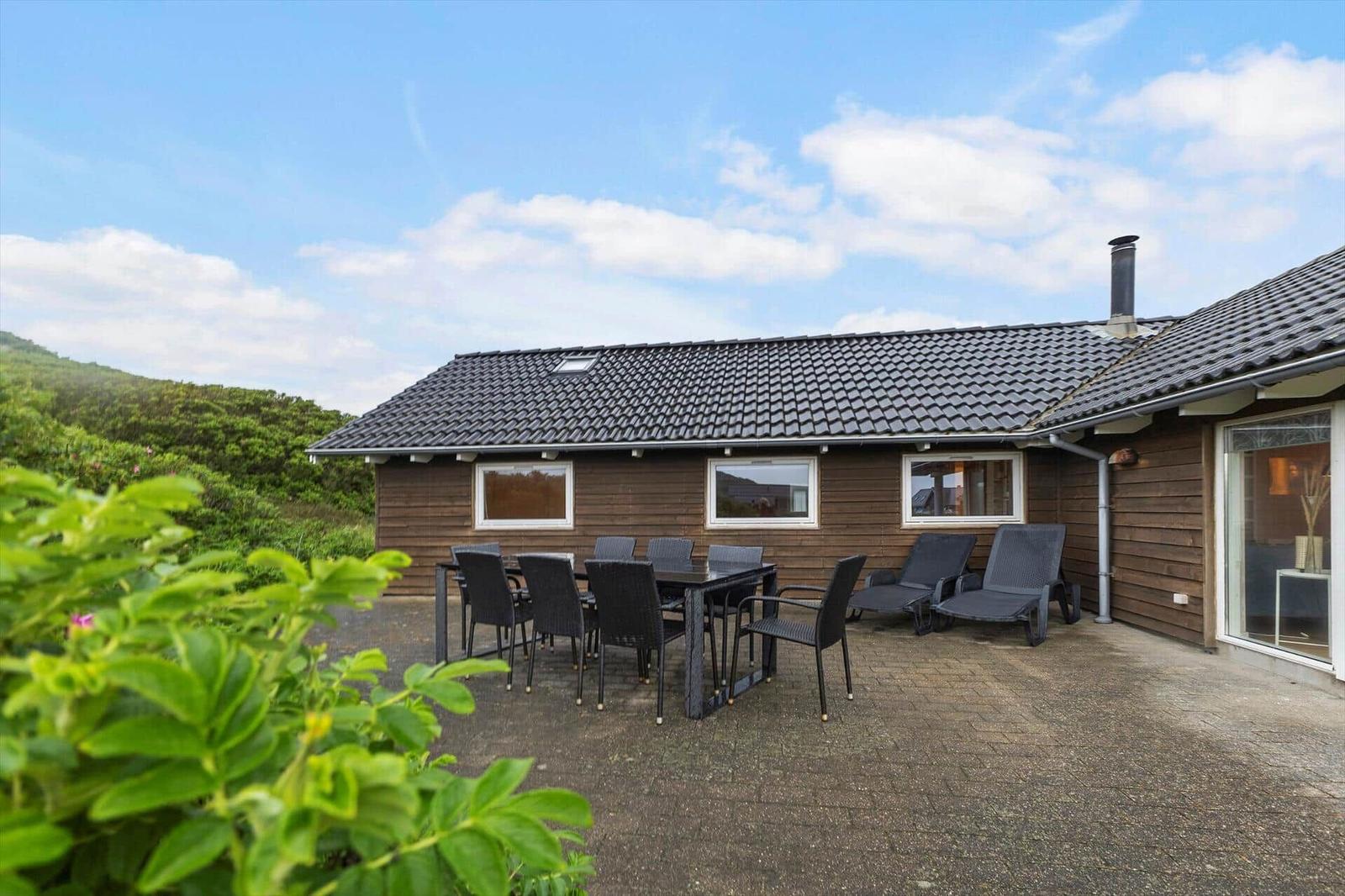 Holzhaus mit Terrasse, Gartenmöbeln und Blick auf grüne Hügel unter blauem Himmel.