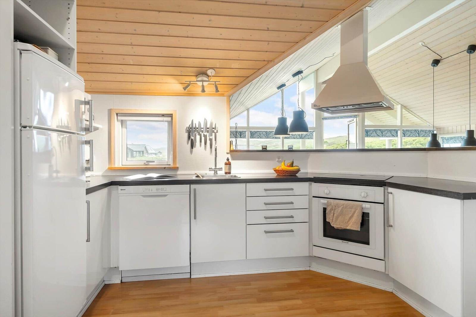 Küche mit weißen Schränken, dunkler Arbeitsplatte und Holzdecke. Fenster bieten Blick auf Landschaft.