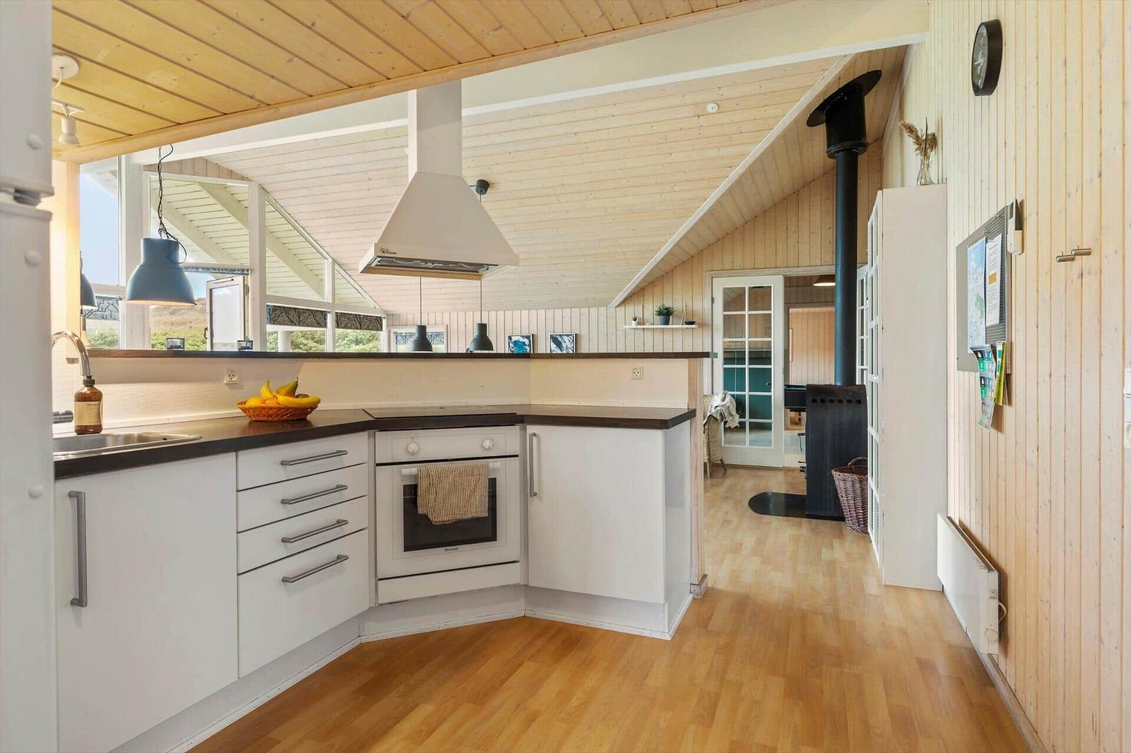 Küche mit weißen Schränken, dunklem Arbeitsplatz und Holzboden. Im Hintergrund ist ein Ofen und eine Tür zu sehen.