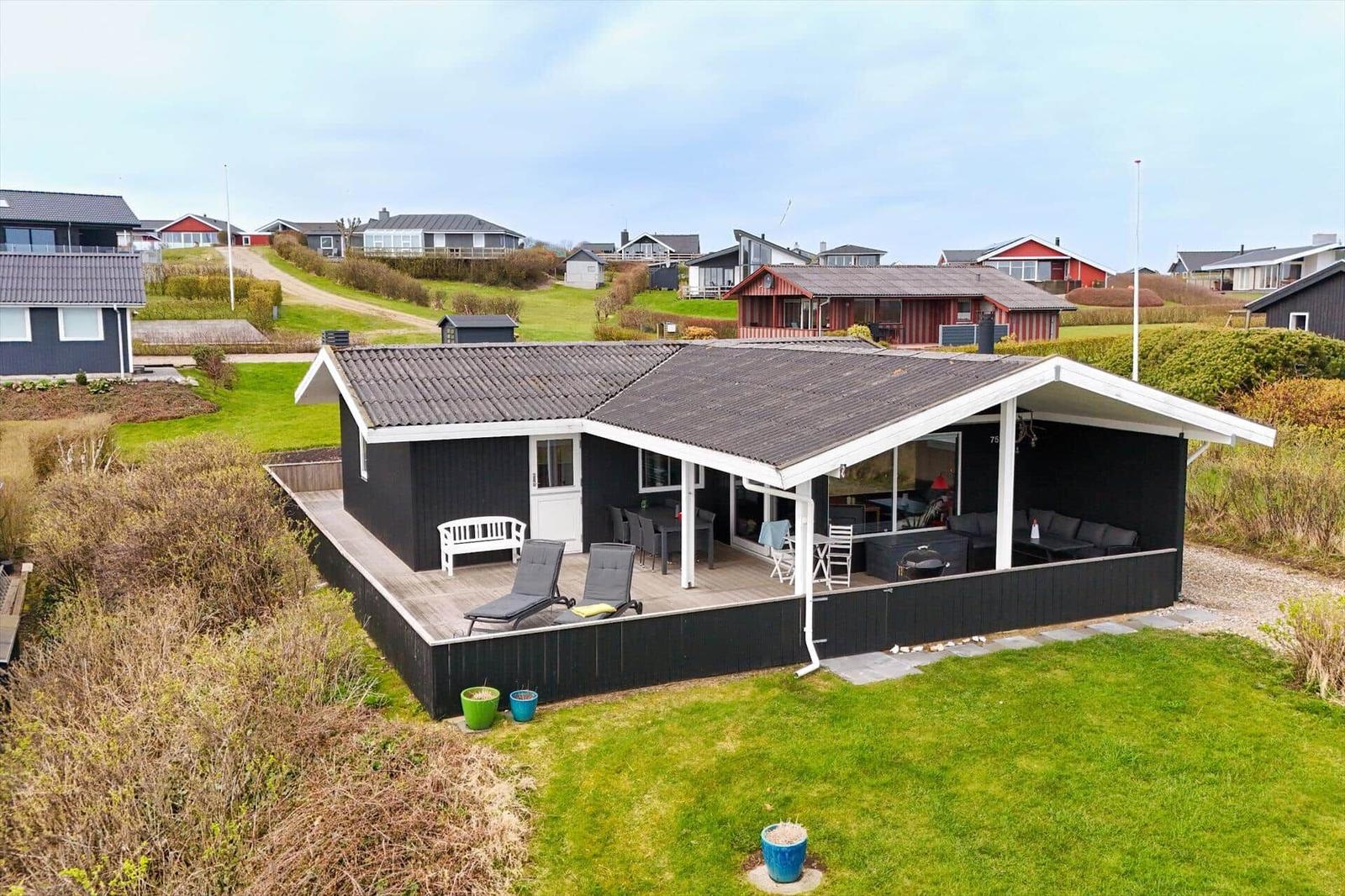 Schwarzes Ferienhaus mit Terrasse, Liegen und Garten. Blick auf weitere Häuser im Hintergrund.