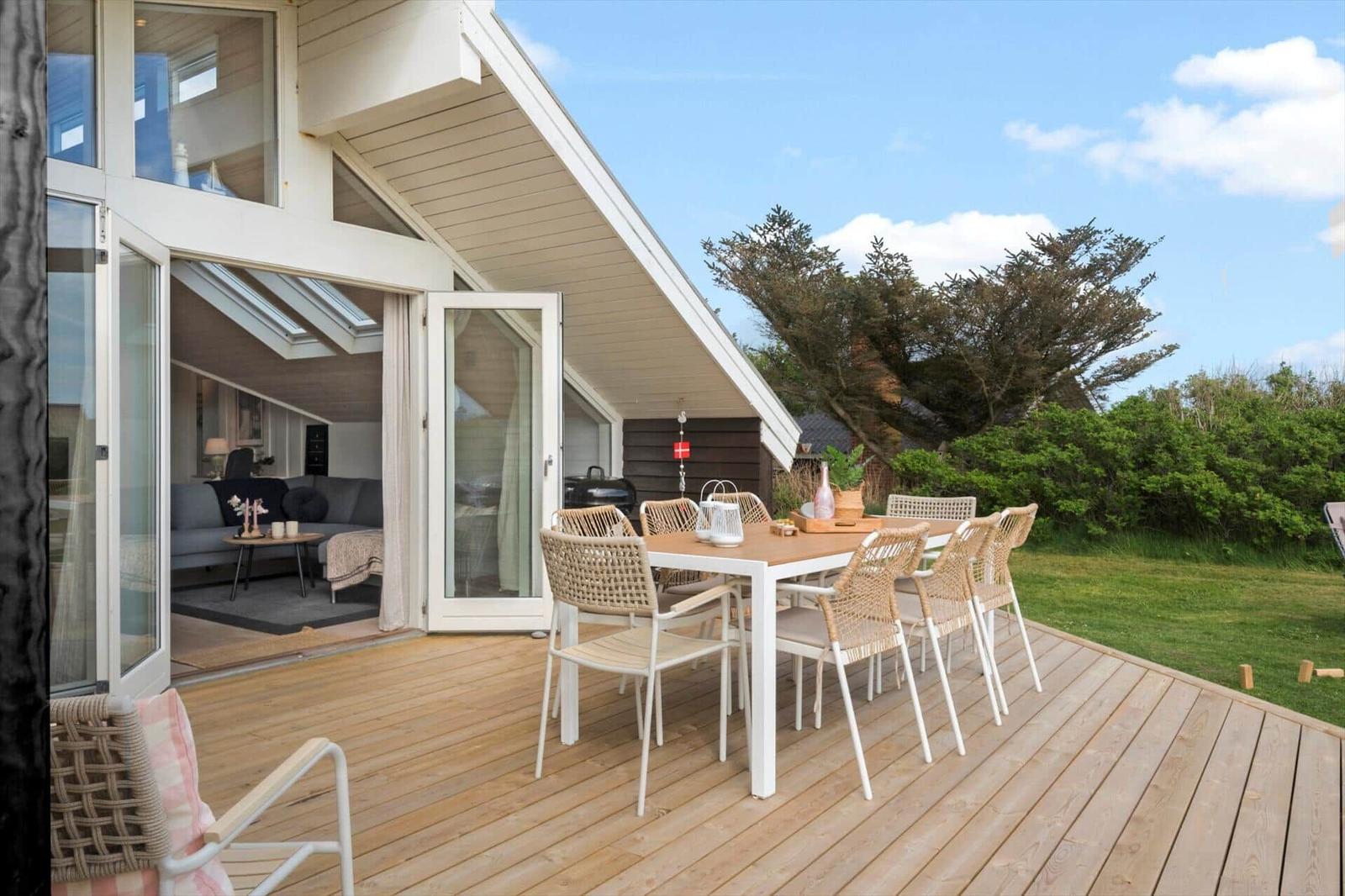 Terrasse mit Esstisch und Stühlen. Durchgehendes Glas führt ins Innere. Grün hinter der Terrasse.