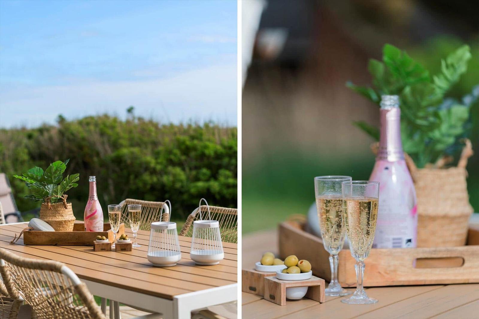 Außenbereich mit Tisch, Stühlen, Flasche und Gläsern. Grüne Vegetation im Hintergrund.