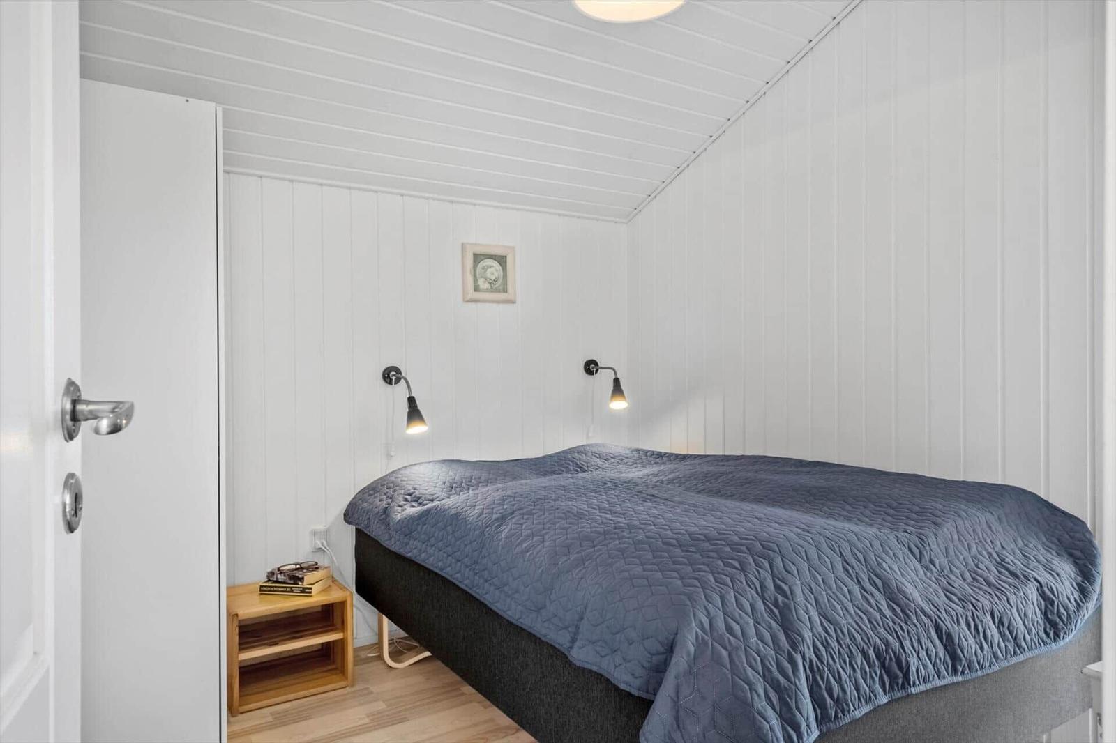 Schlafzimmer mit Bett, Nachttisch und Wandlampen. Wände und Decke sind weiß verkleidet.