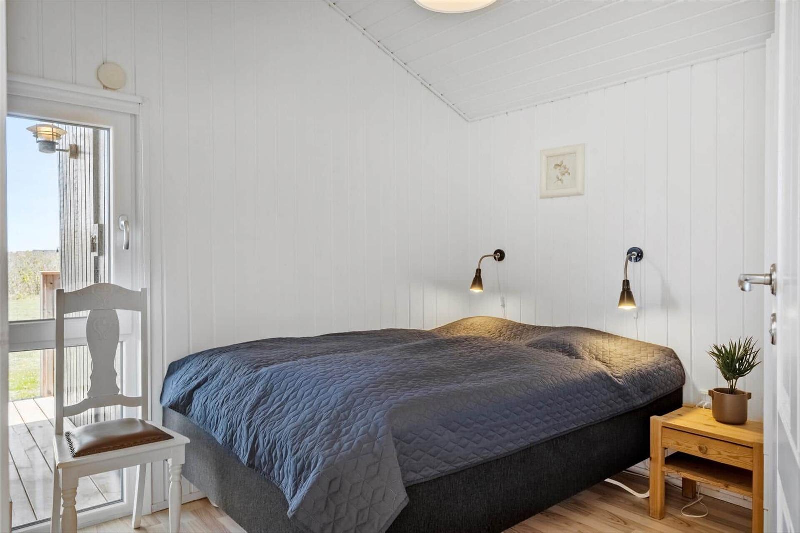 Schlafzimmer mit Bett, Nachttisch und Stuhl. Weiße Wände, Holzboden und Fenster zum Balkon.