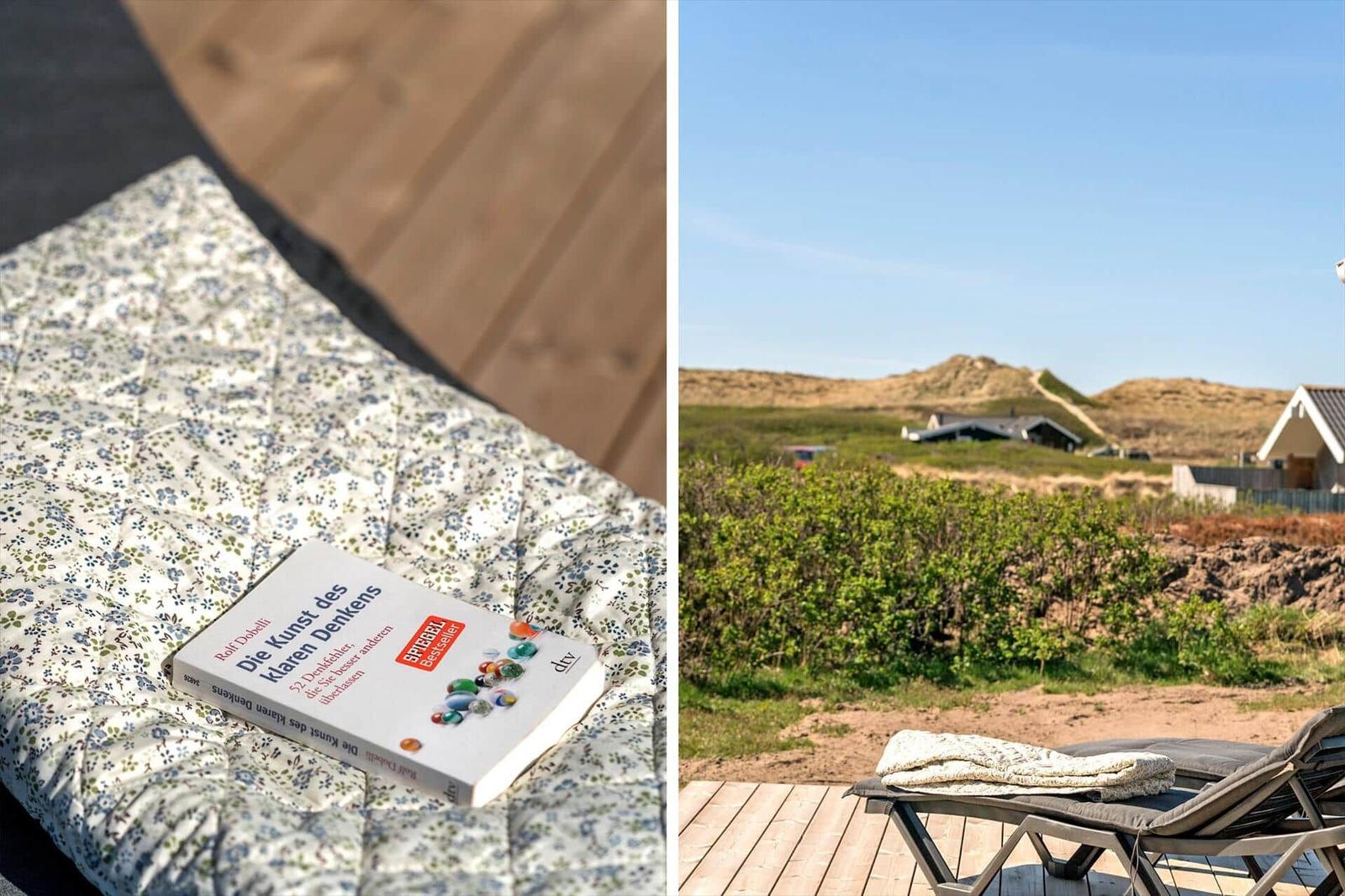 Ein Buch liegt auf einem gemusterten Kissen. Im Hintergrund eine Terrasse mit Blick auf Dünen.