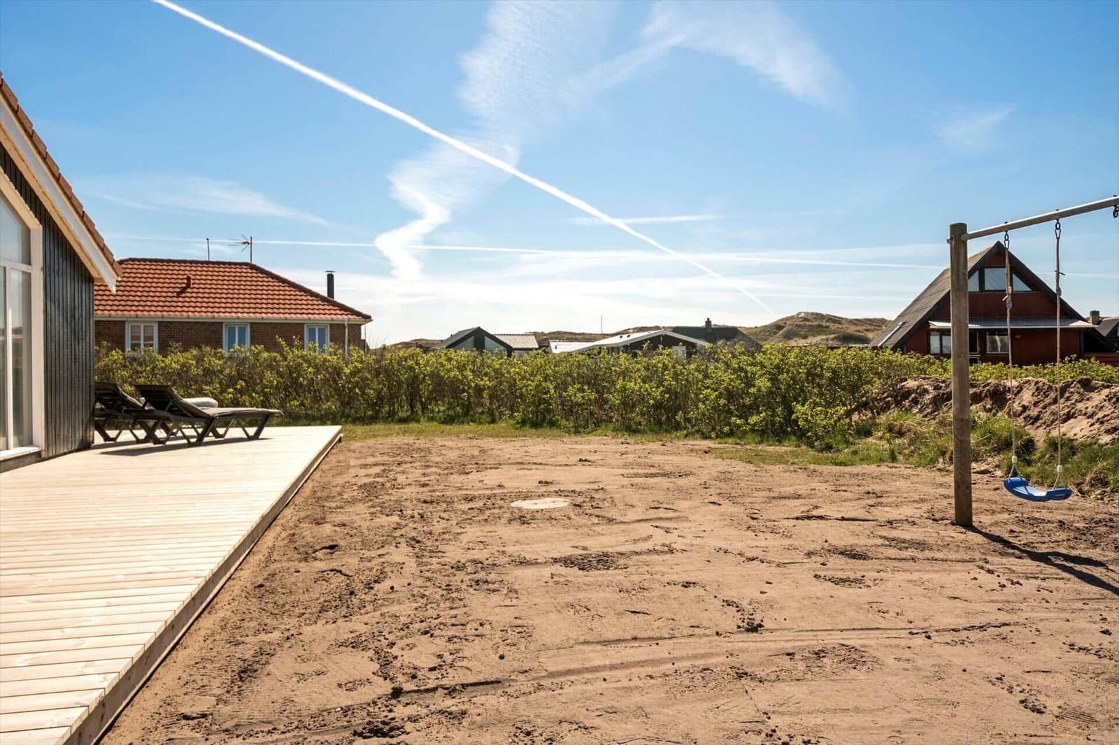 Terrasse mit Liegestühlen und Sandfläche. Hintergrund: Grünhecke und Häuser.