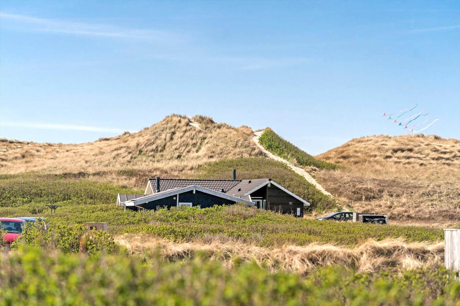 Haus mit Dach und Schornstein zwischen Dünen. Mehrere Kites fliegen im Himmel.