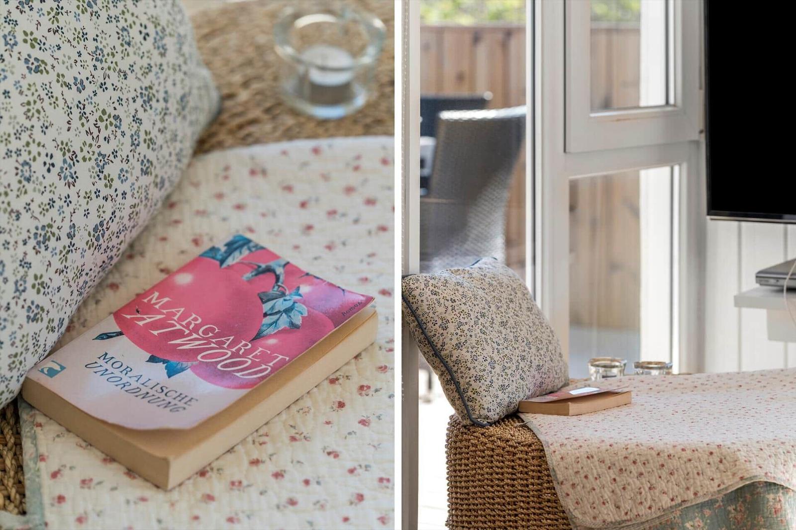 Ein Buch liegt auf einem floralen Kissen. Durch ein Fenster ist ein Balkon mit Stuhl sichtbar.