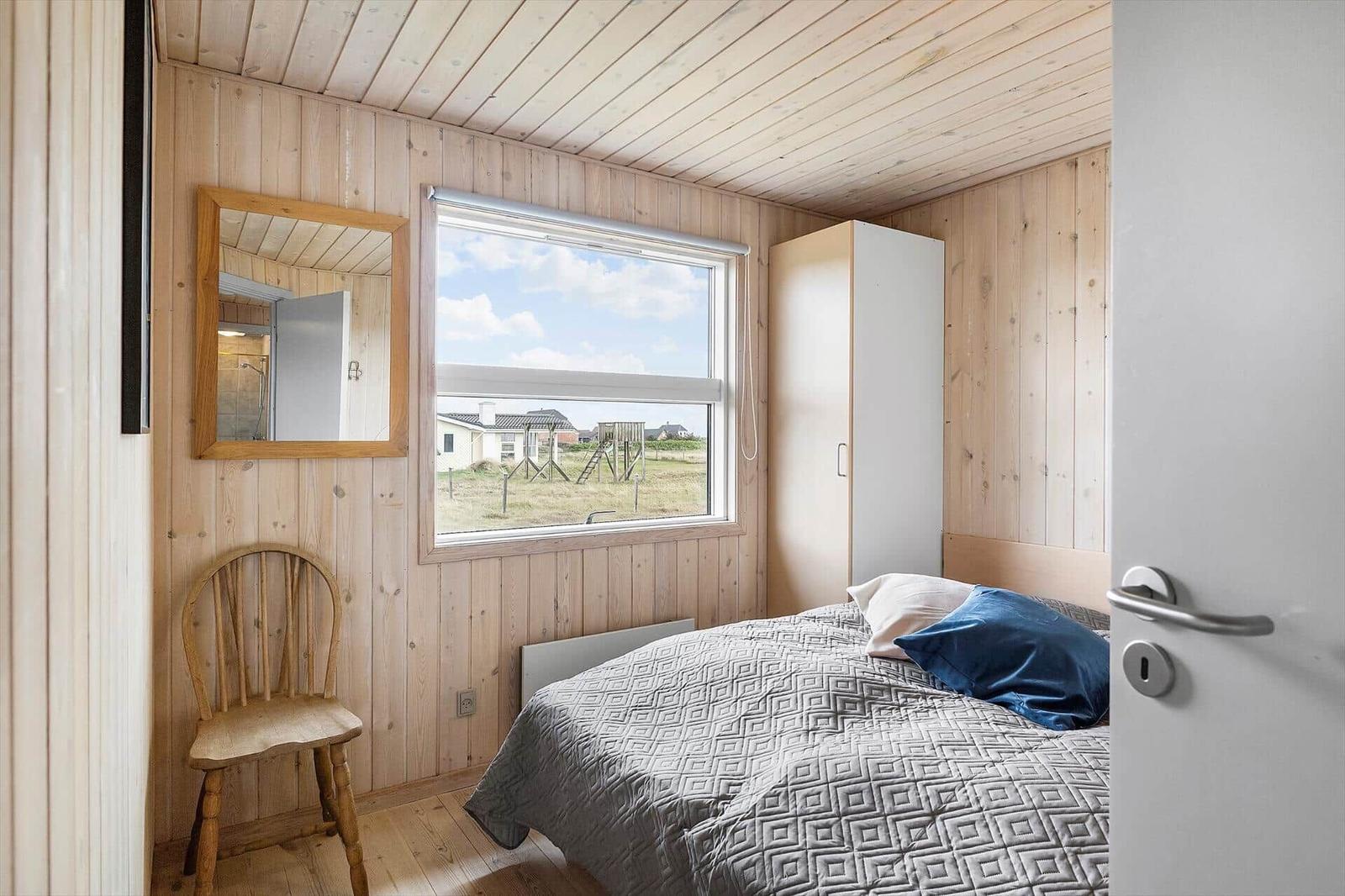 Schlafzimmer mit Holzwand, Bett, Stuhl und Fensterblick auf die Landschaft.