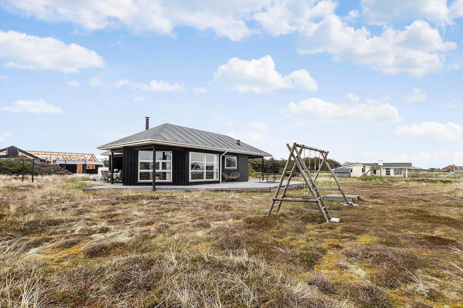 Schwarzes Haus mit Holzterrasse und Schaukel im Grasland unter blauem Himmel.
