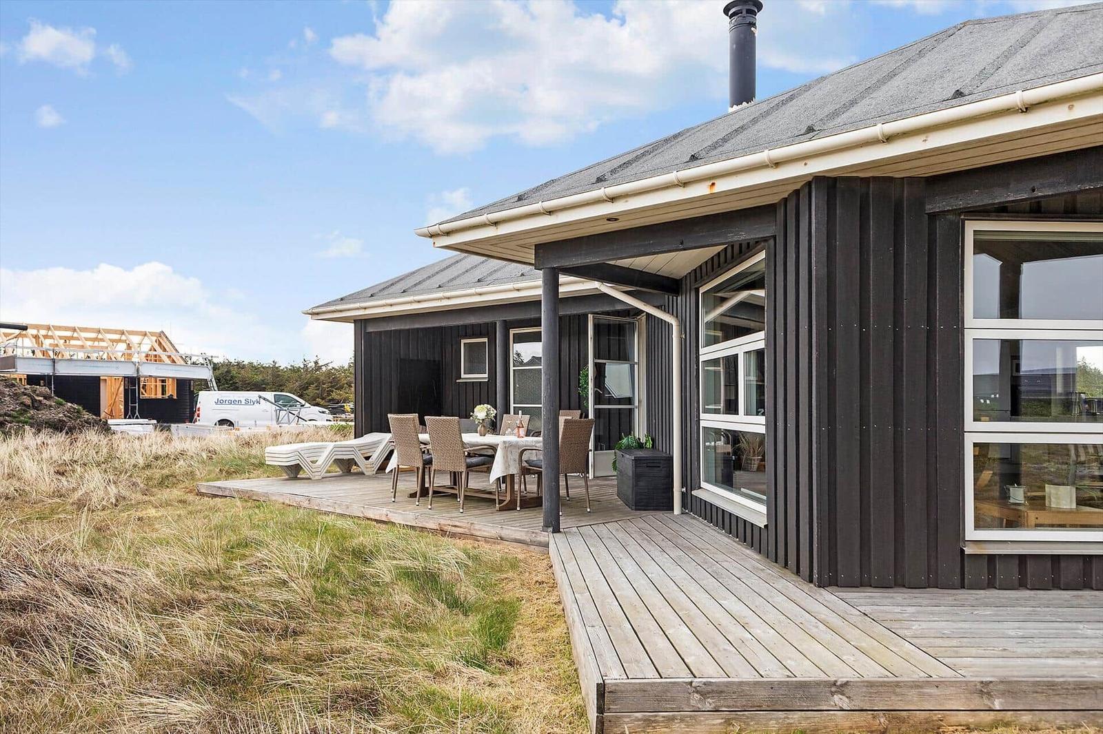 Moderne Holzhütte mit Terrasse und Gartenmöbeln. Hintergrund: Baustelle und Himmel.