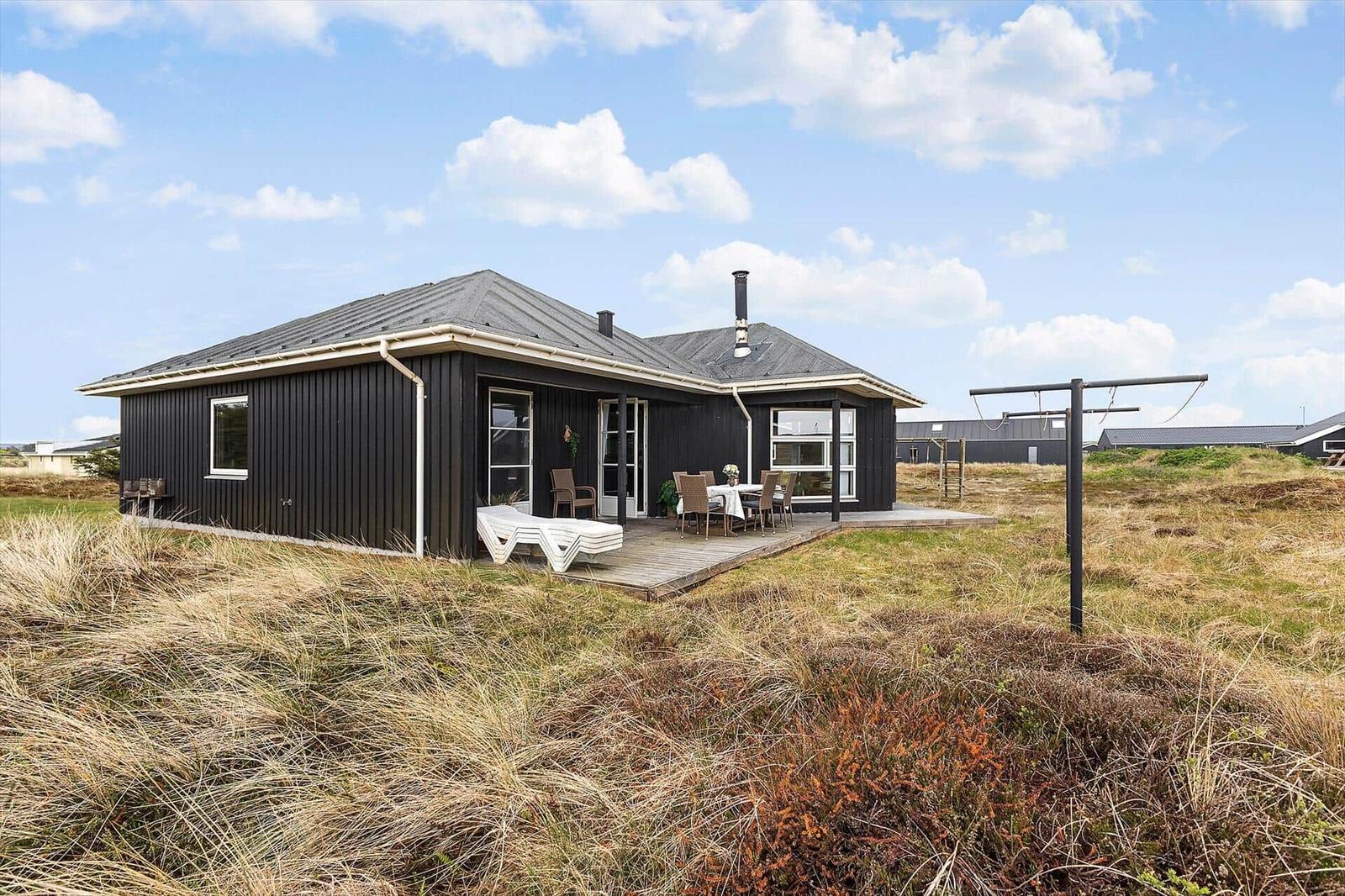 Schwarzes Haus mit Terrasse und Garten. Tisch, Stühle und Liege sind draußen.