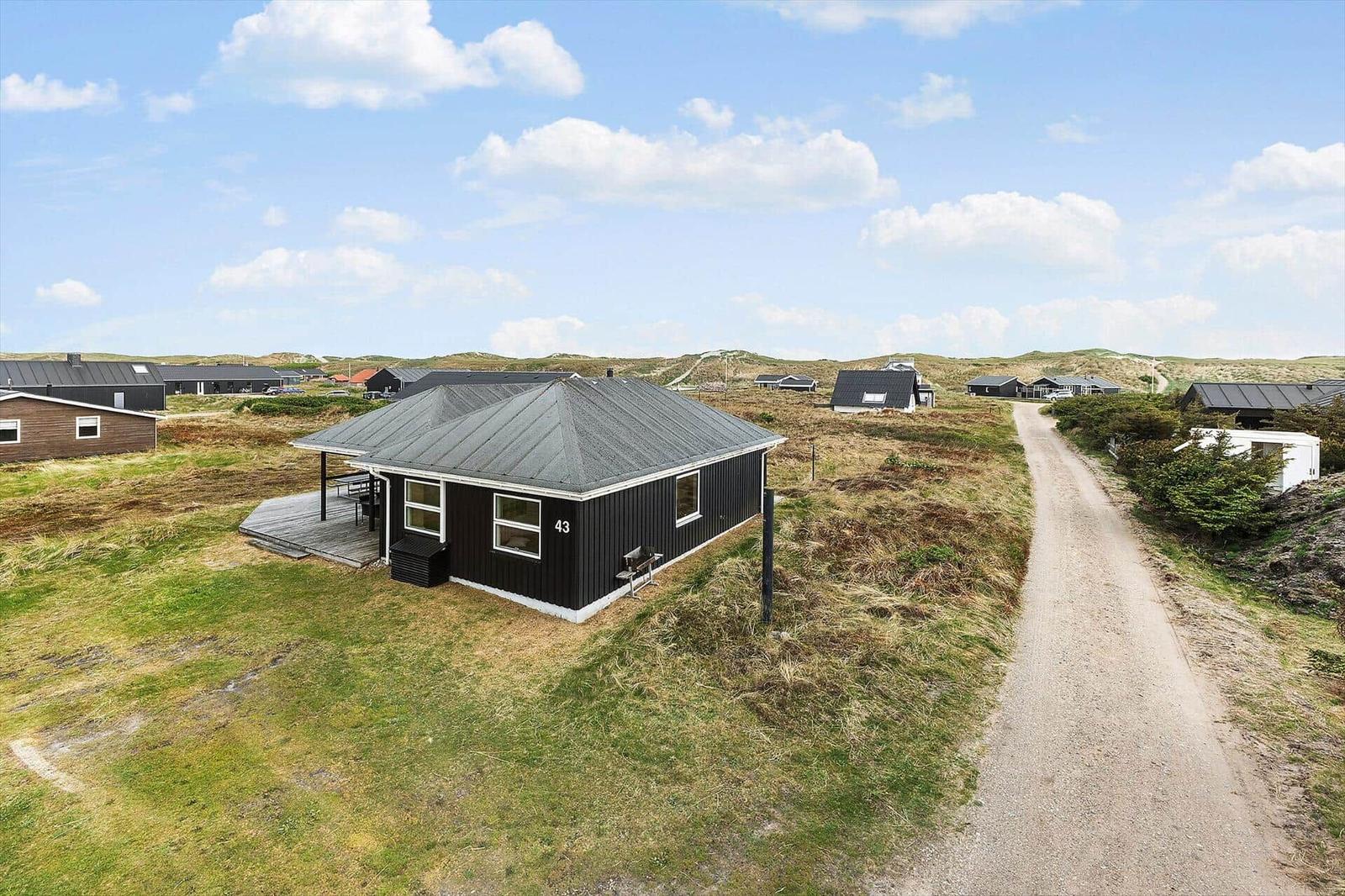 Schwarzes Ferienhaus mit Terrasse, umgeben von Gras und Dünen. Zahlreiche Häuser sind im Hintergrund sichtbar.