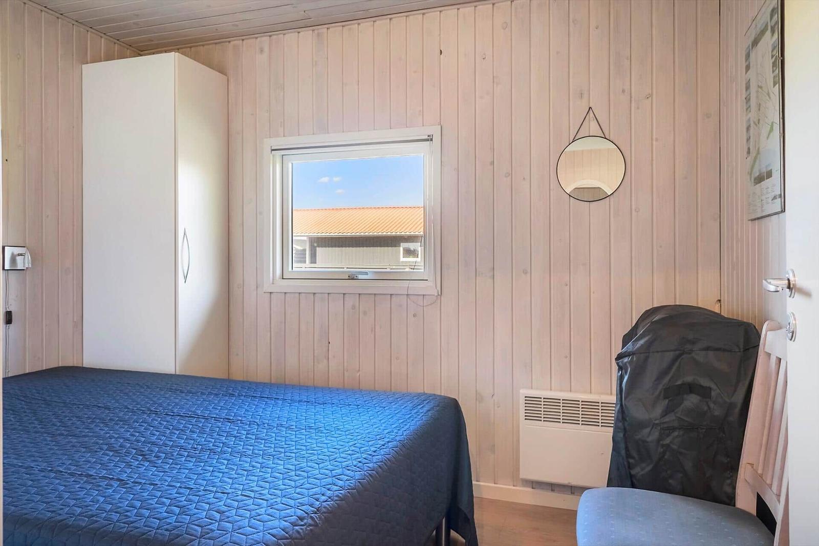 Schlafzimmer mit Bett, Kleiderschrank und Fenster. An der Wand hängt ein runder Spiegel.