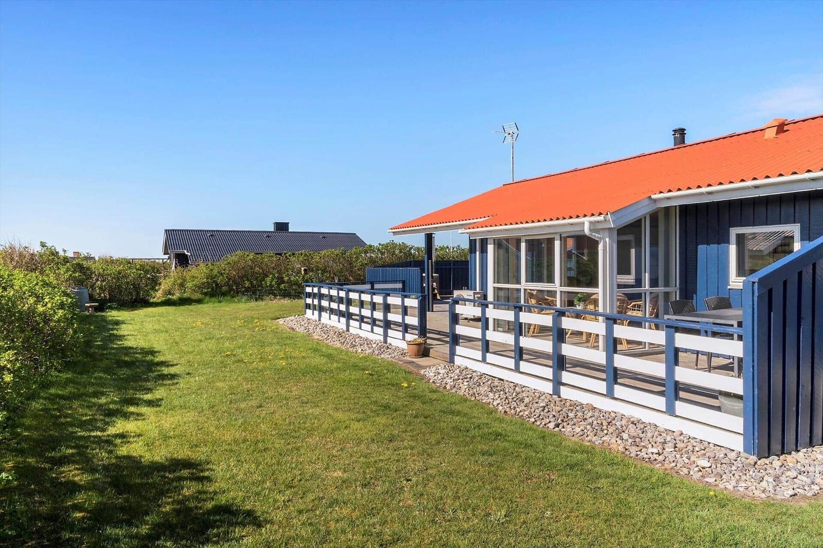 Blau gestrichenes Haus mit rotem Dach und Terrasse. Grasfläche und Zaun im Vordergrund.