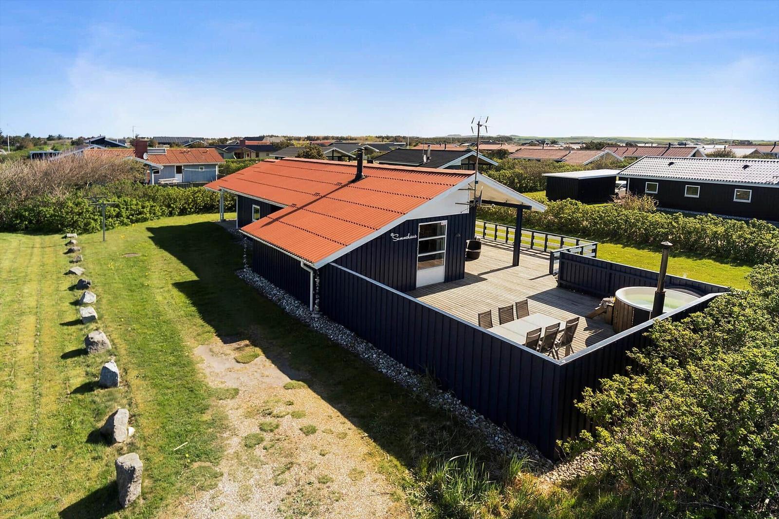Schwarzes Ferienhaus mit Terrasse und Whirlpool. Roter Dachfirst. Grünflächen und Nachbarhäuser im Hintergrund.