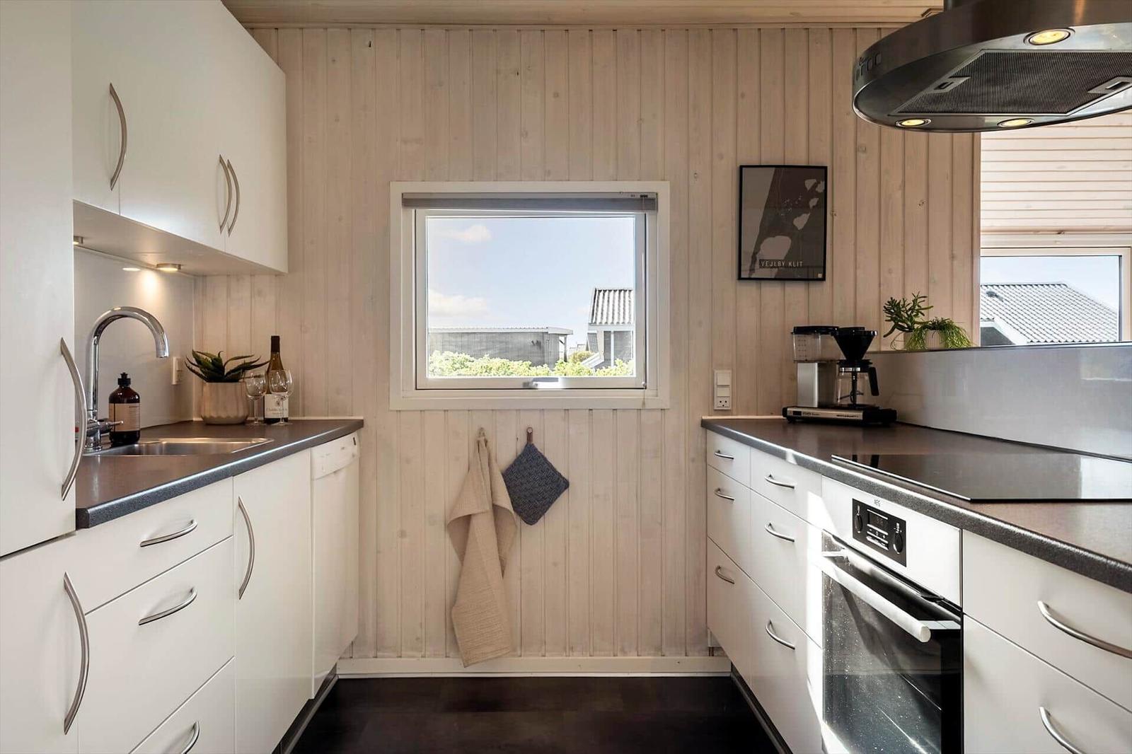 Küche mit weißen Schränken, dunklem Arbeitsplatz und Holzwand. Fenster mit Blick nach draußen.