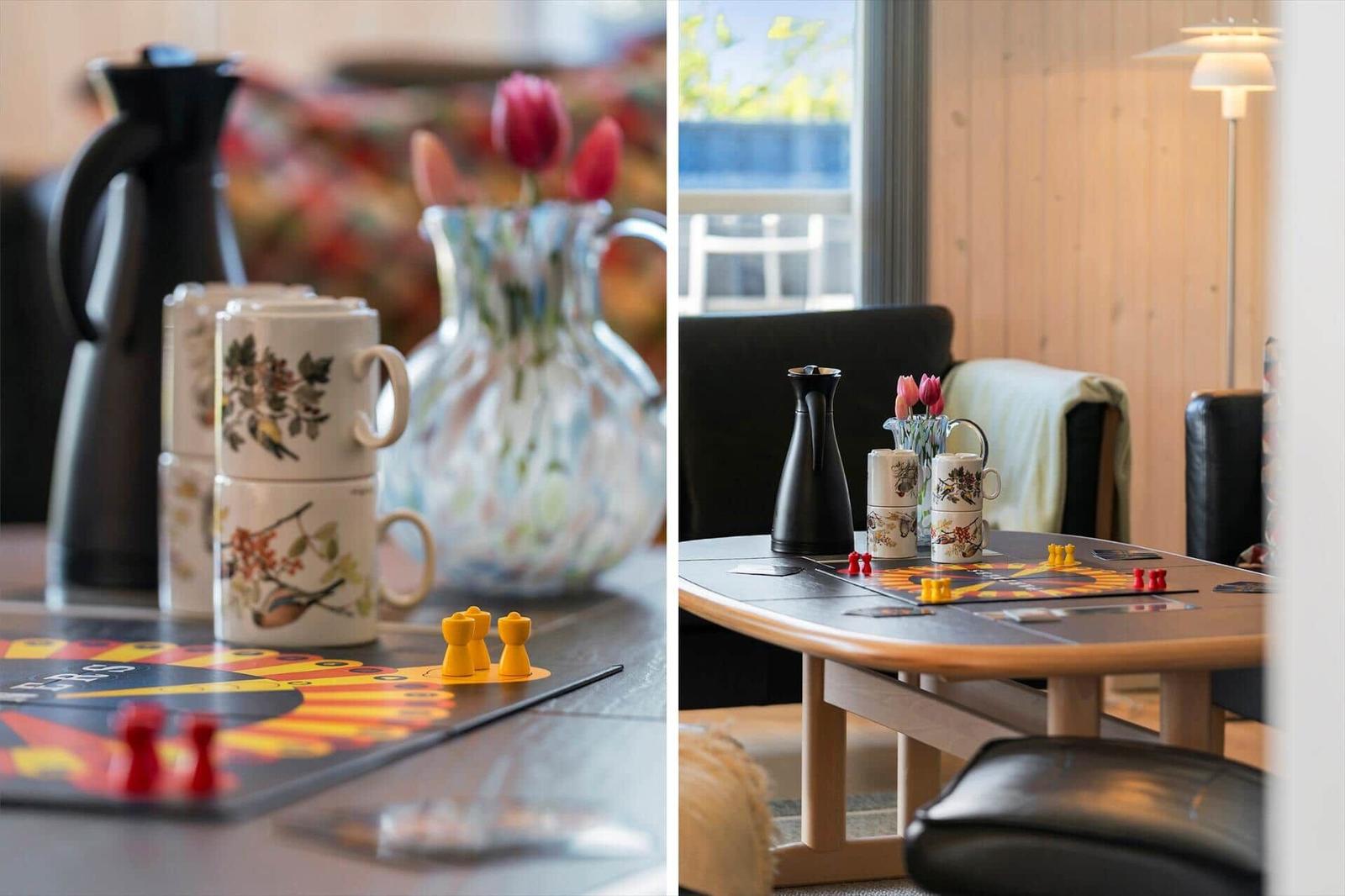 Tisch mit Spielplatte, Tassen und Blumen in einem Wohnzimmer mit Holzwand und Sitzgelegenheiten.