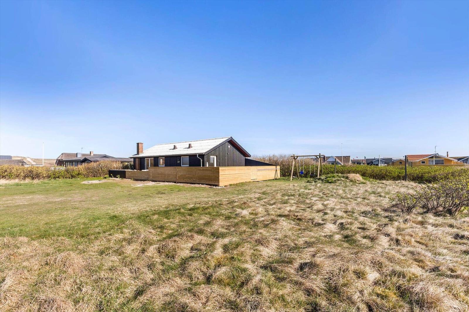 Ein Haus mit Holzterrasse und Spielplatz auf einer grünen Wiese unter blauem Himmel.