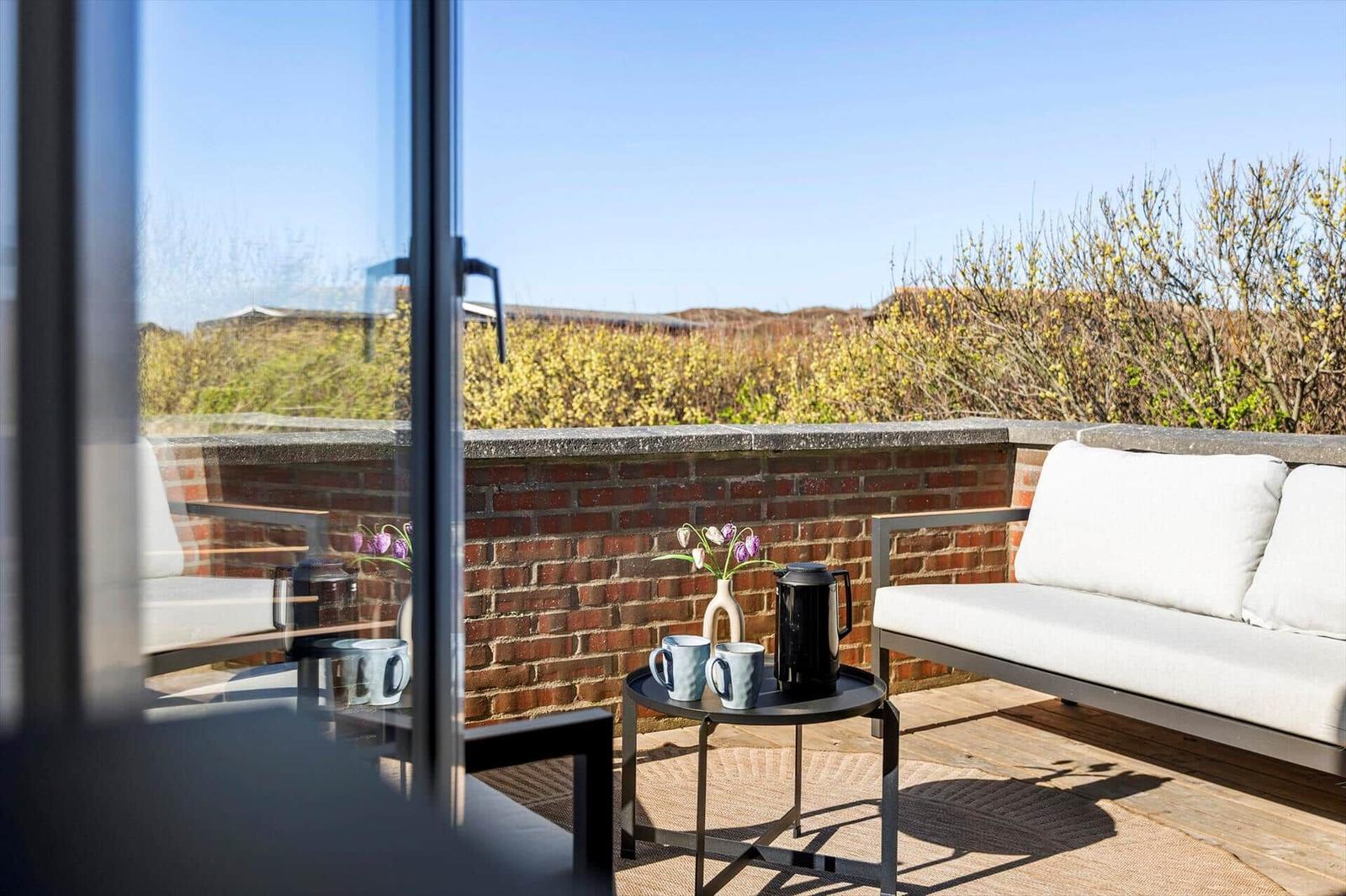 Terrasse mit Sofa, Tisch und Blumen. Blick auf grüne Hecke und Himmel.