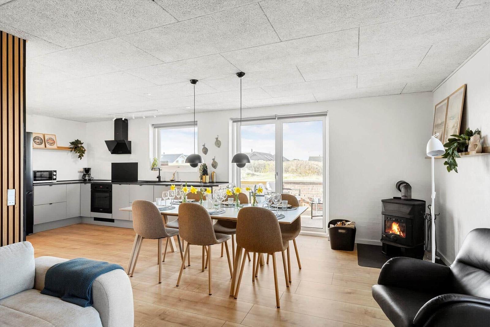 Offener Raum mit Küchenzeile, Esstisch und Kaminofen. Holzboden und große Fenster.