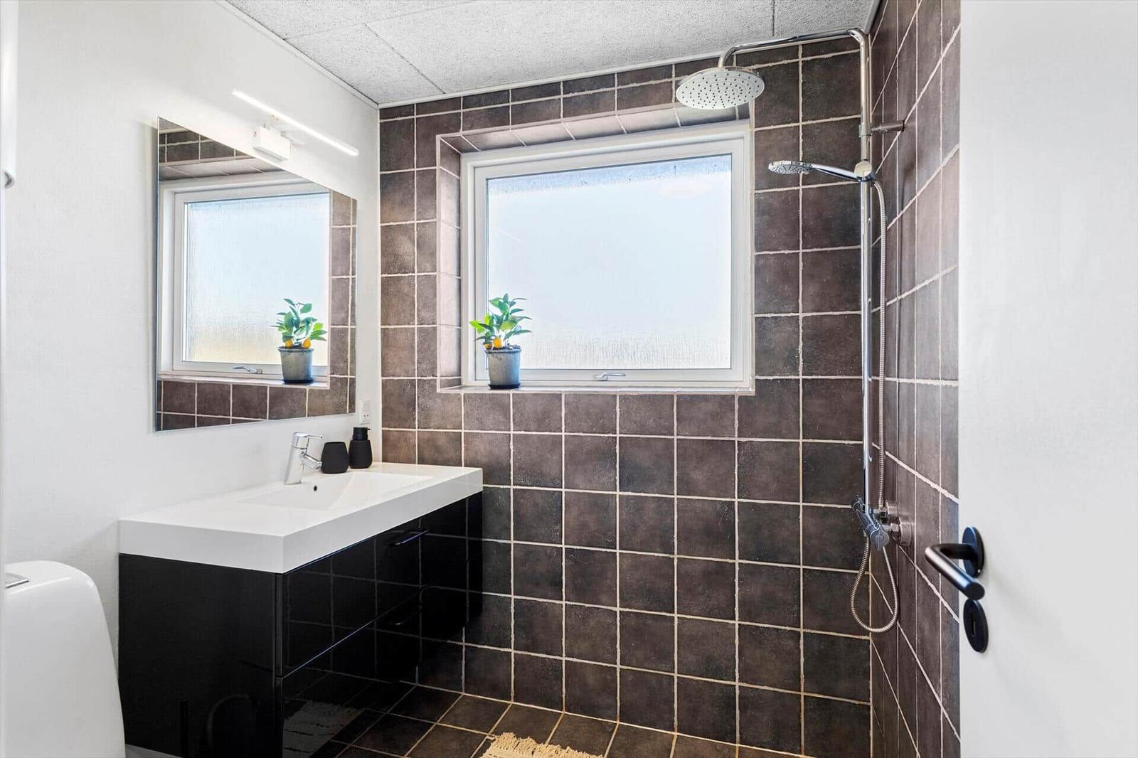 Bad mit Dusche, Waschbecken und Fenster. Wände und Boden sind mit dunklen Fliesen verkleidet.