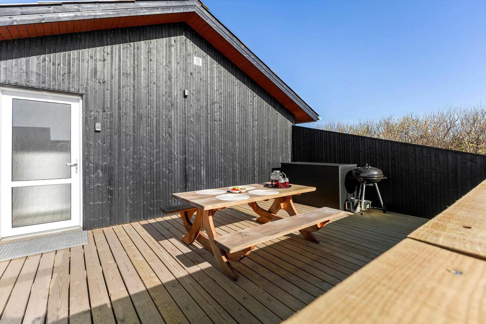 Terrasse mit Holztisch, Bänken und Grill vor einem Haus mit dunkler Holzfassade.