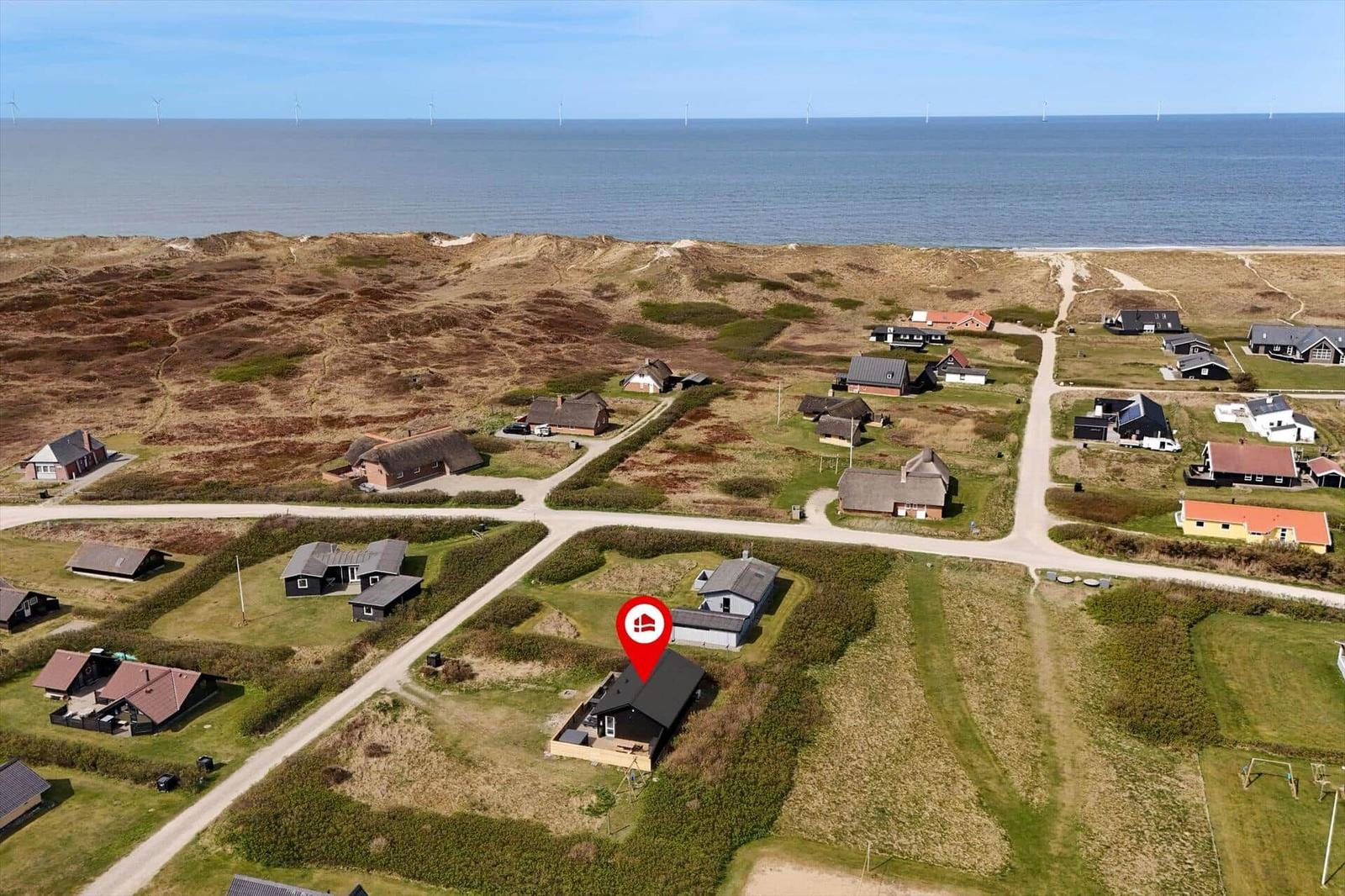 Aerielansicht einer Ferienwohnung in einer Küstenlandschaft mit Häusern und Windrädern am Horizont.