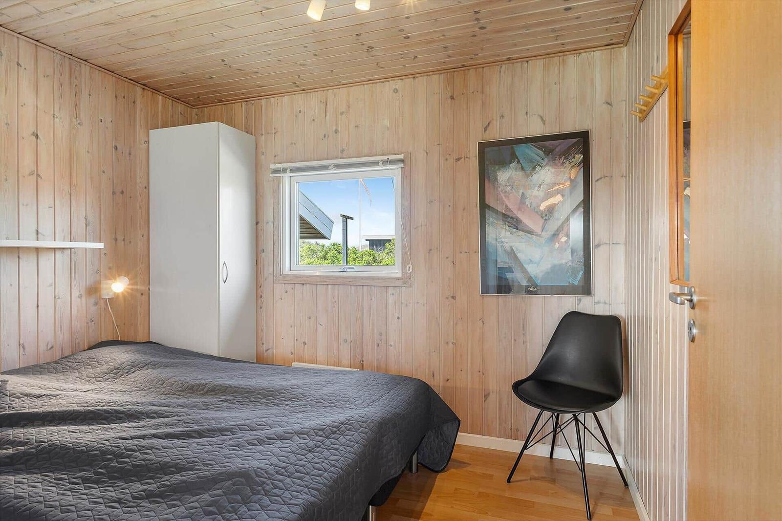 Schlafzimmer mit Holzwand, Bett, Schrank, Stuhl und Fenster.