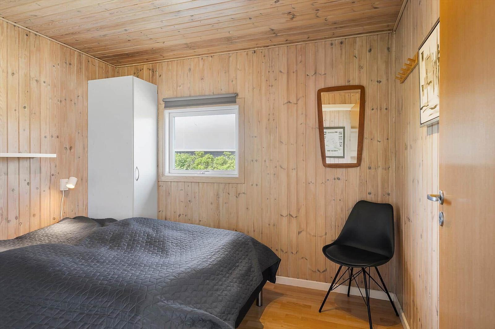 Schlafzimmer mit Holzwand, Bett, Schrank, Stuhl und Fenster.