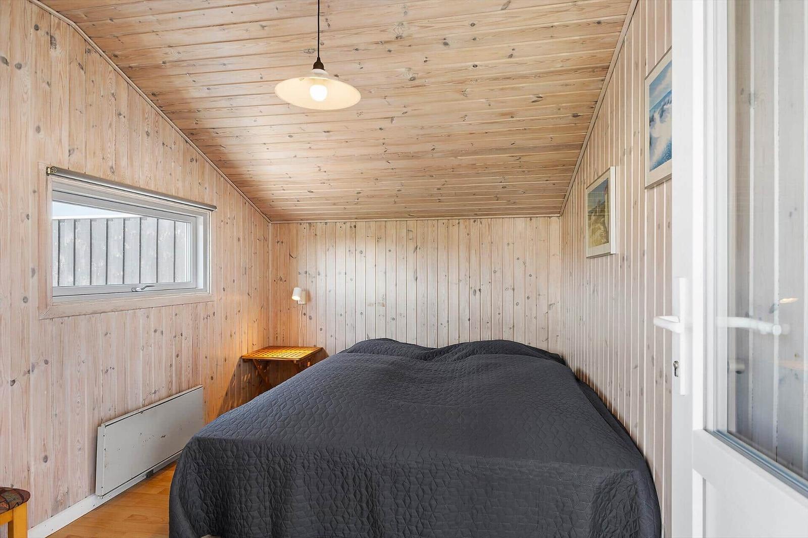 Schlafzimmer mit Holzwandverkleidung, Bett mit dunkler Bettwäsche und Fenster.