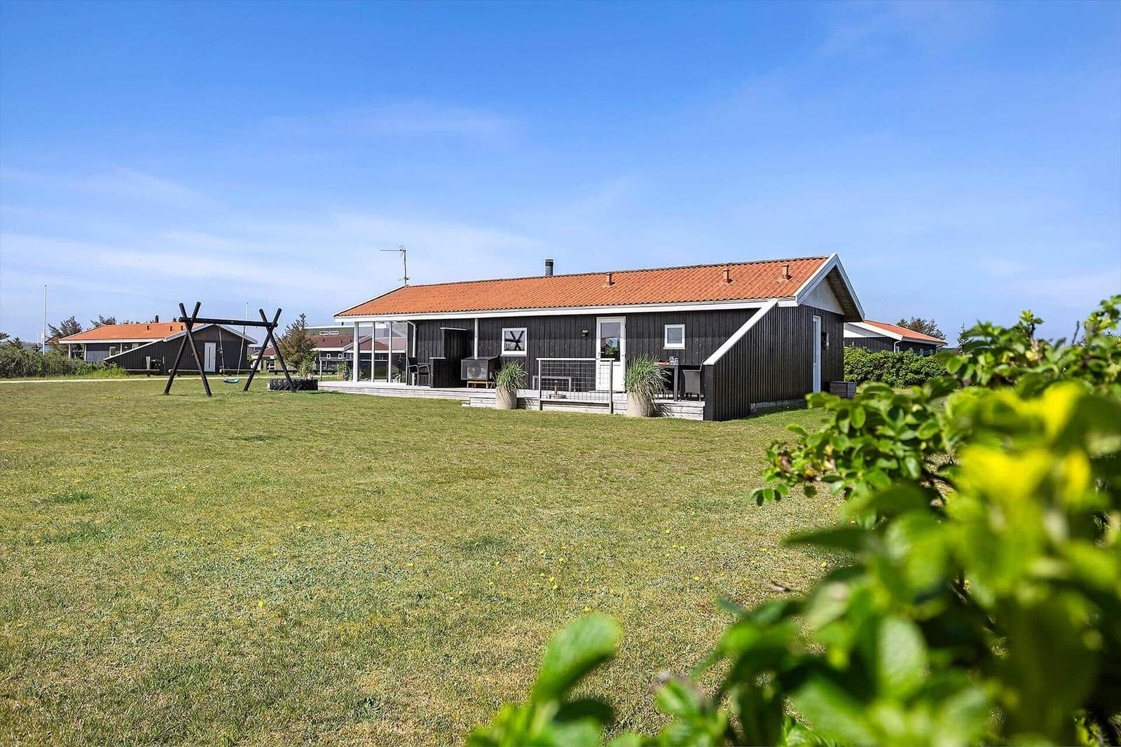 Ein Haus mit rotem Dach und Holzfassade steht auf einer grünen Wiese. Eine Terrasse und ein Spielplatz sind sichtbar.