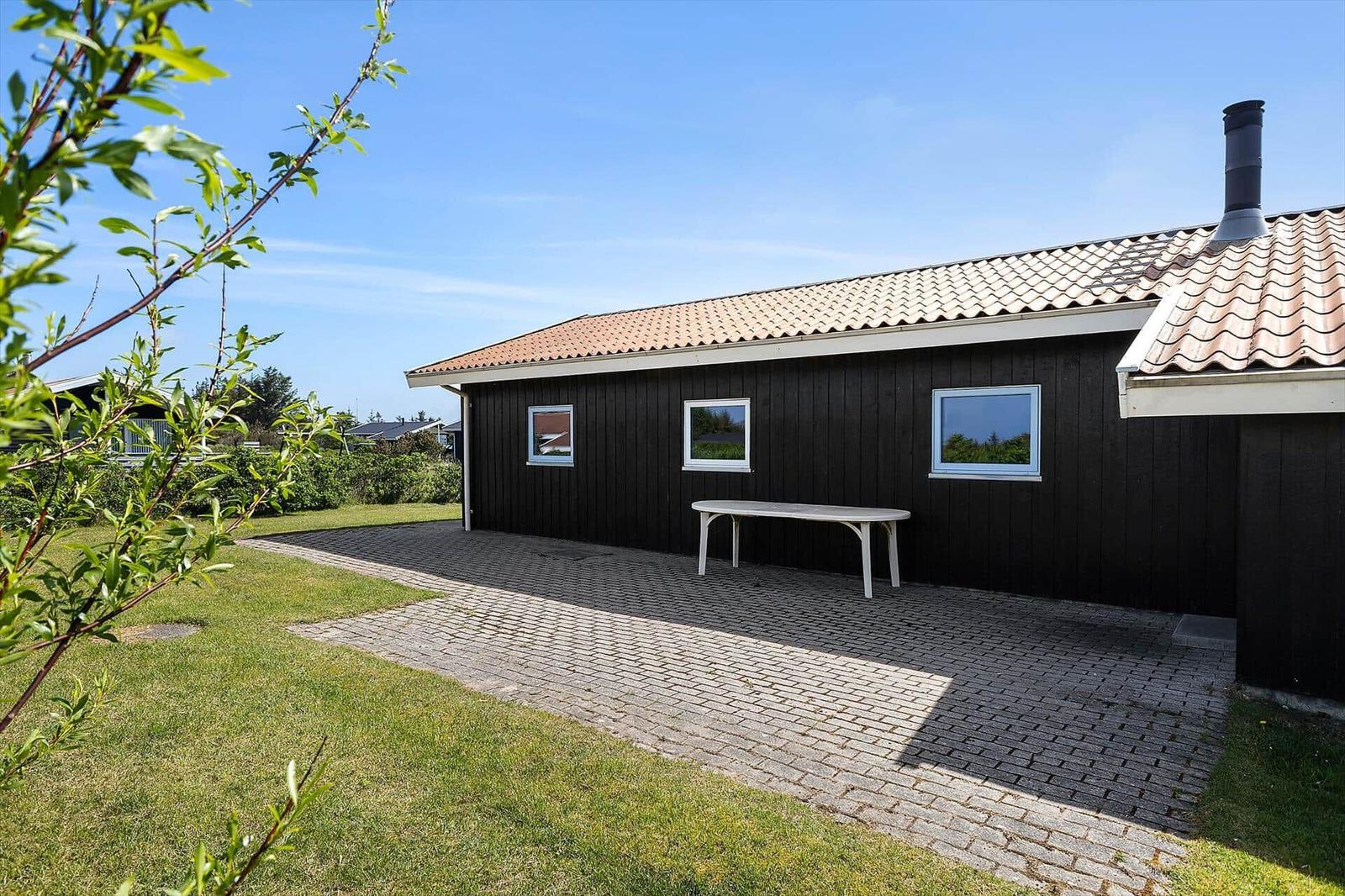 Ein dunkles Holzhaus mit Terrasse, Tisch und Grünfläche unter blauem Himmel.