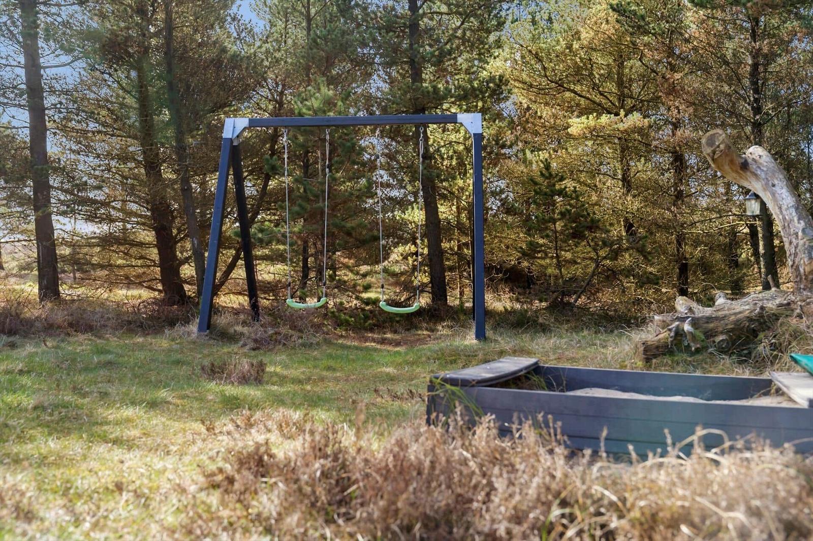 Schaukel und Sandkasten im Wald. Gras und Bäume umgeben den Spielplatz.