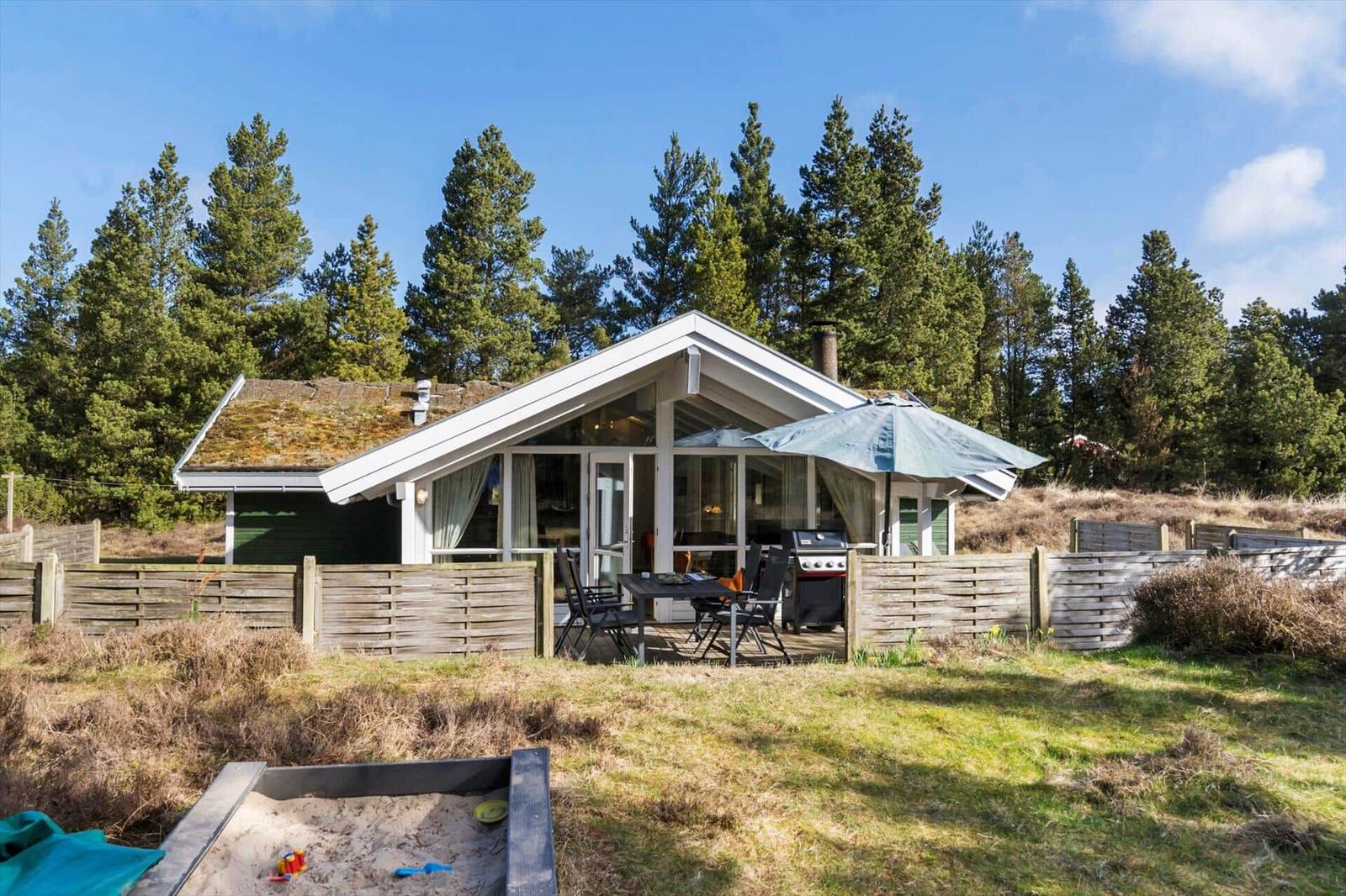 Ein Haus mit Terrasse, Grill und Sandkasten vor einem Wald.