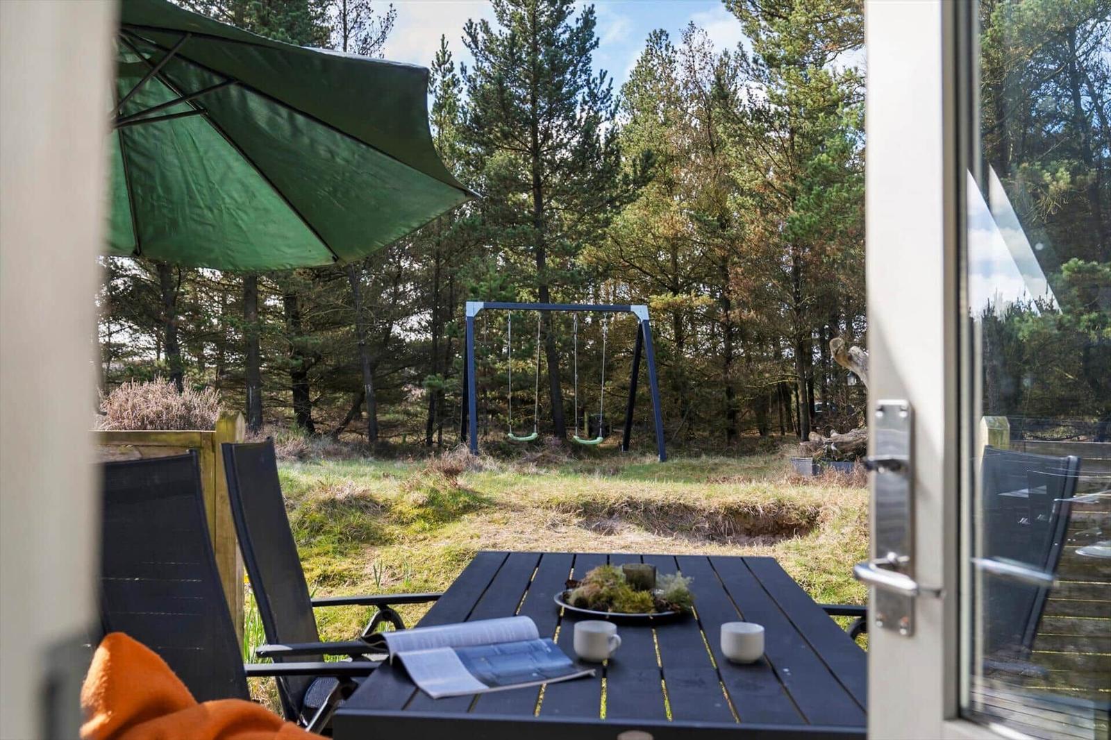 Terrasse mit Tisch, Stühlen und grünem Sonnenschirm. Hintergrund: Wald mit Schaukel.