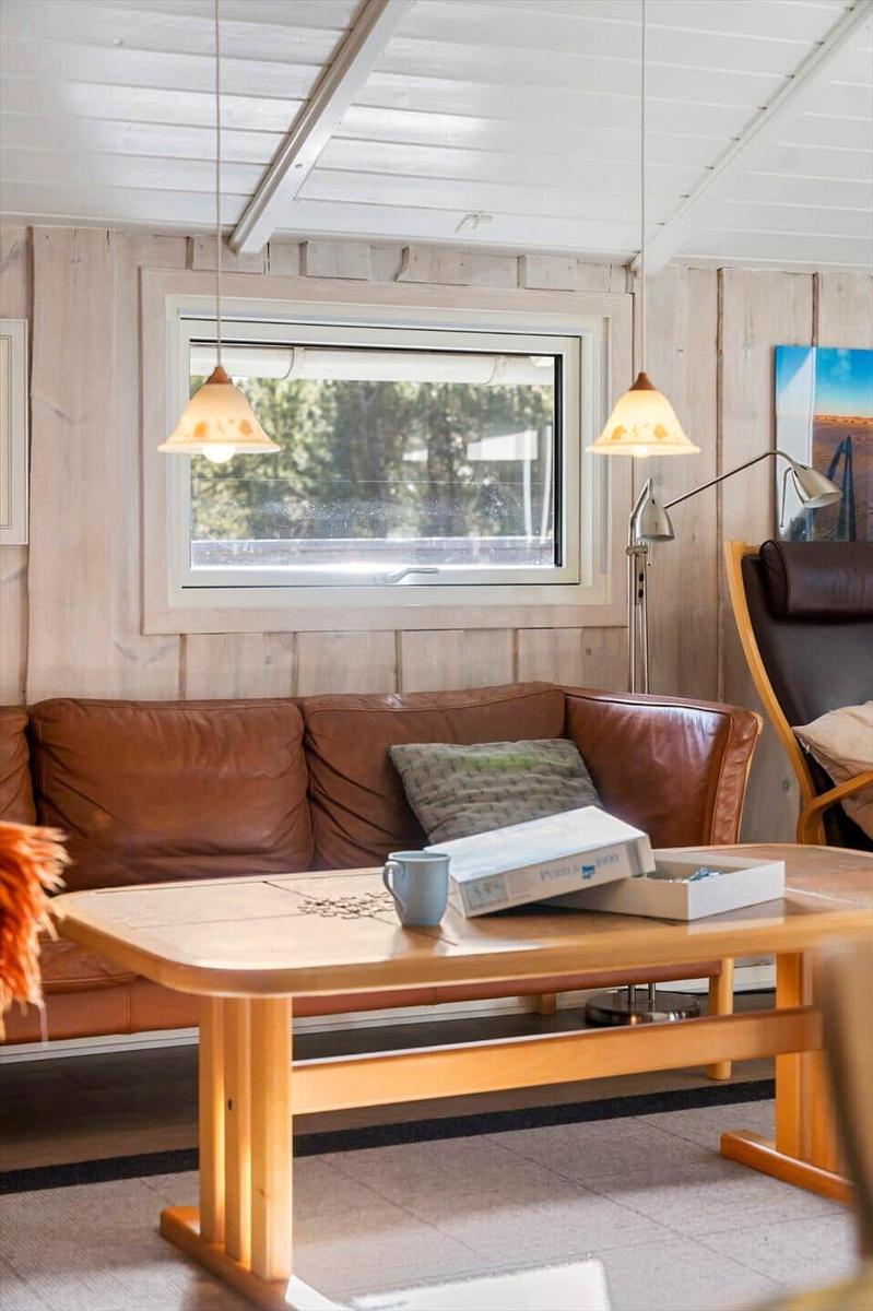 Ein Wohnzimmer mit braunem Sofa, Holztisch und Fenster. Hängelampen und Wandbild sind sichtbar.