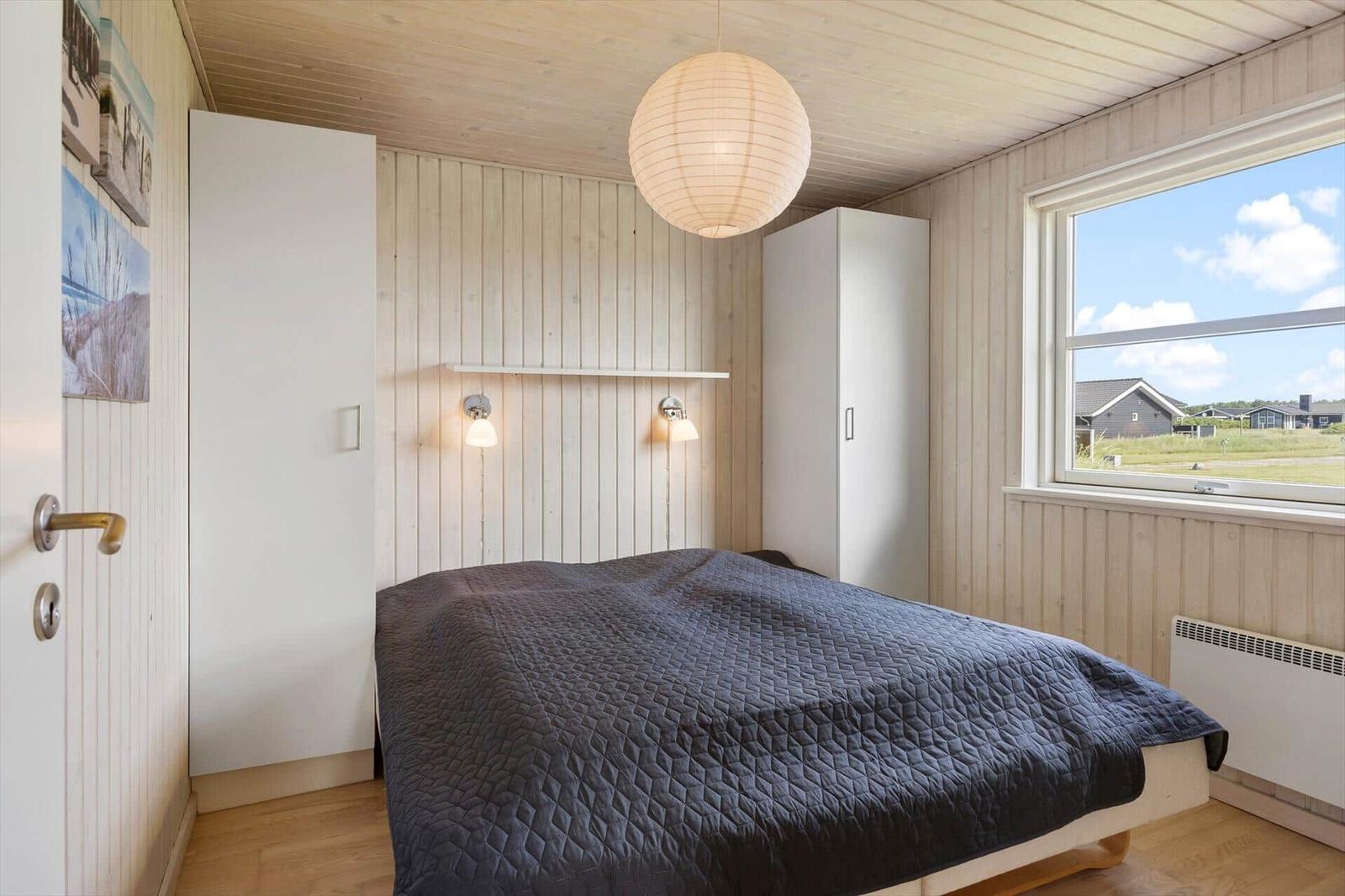 Schlafzimmer mit Bett, weißen Schränken und Holzwand. Fenster zeigt Landschaft.