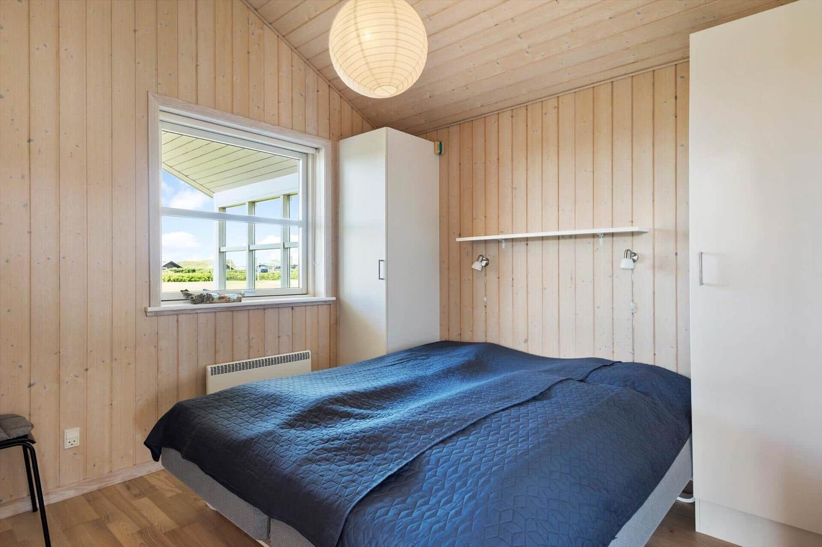 Schlafzimmer mit Holzwand, Bett mit blauer Bettdecke und Fenster mit Blick nach draußen.