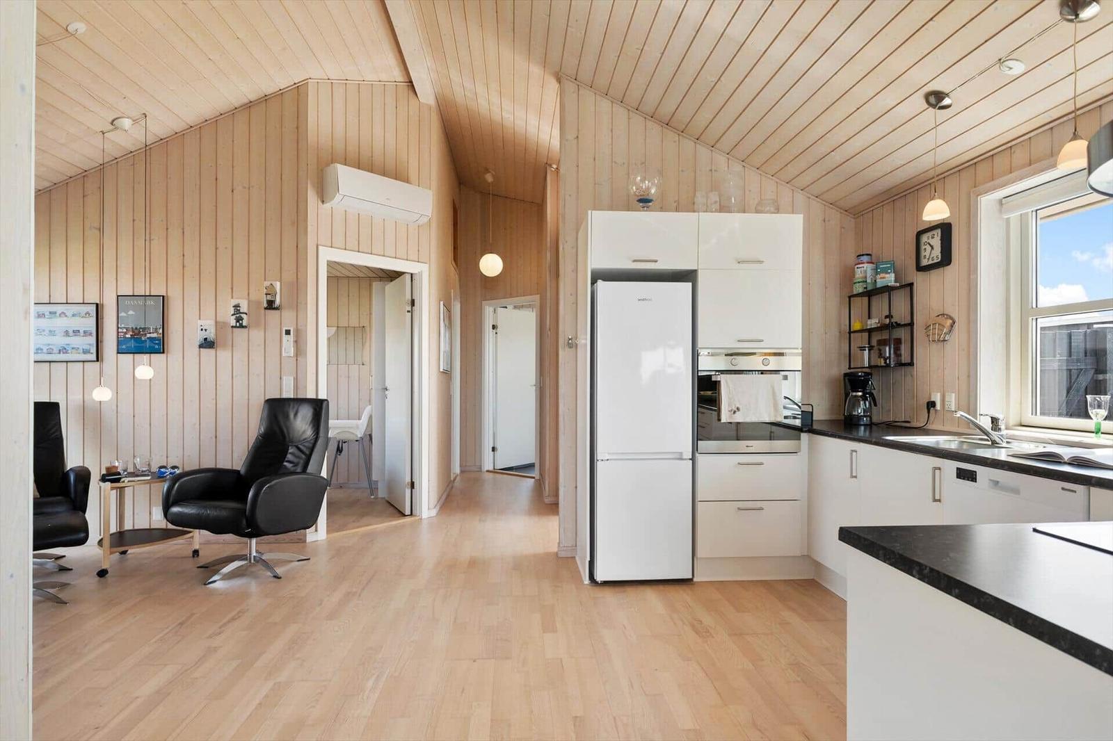 Küche mit weißer Küchenzeile, Kühlschrank und Fenster. Wohnbereich mit zwei schwarzen Sesseln.