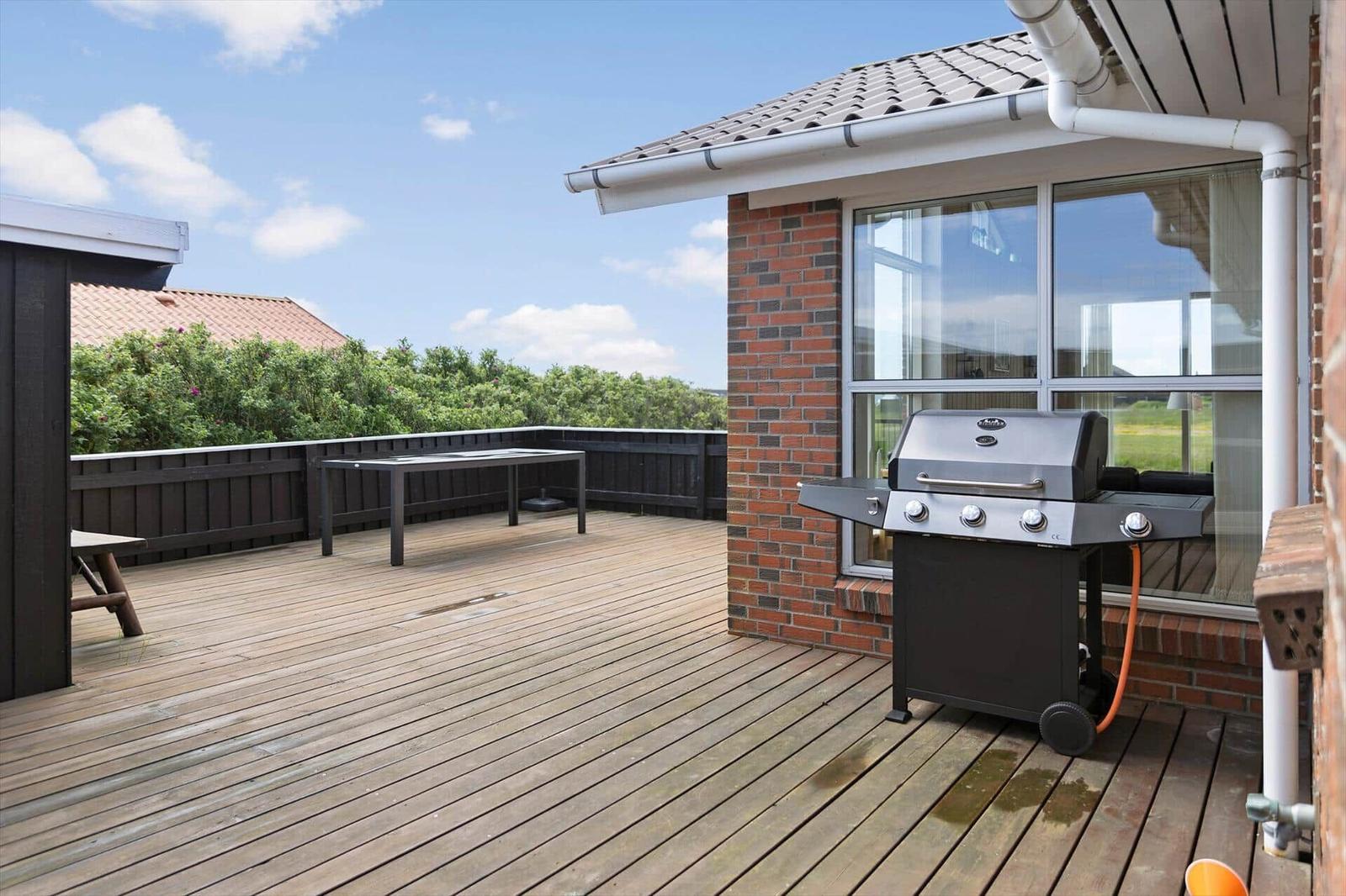 Terrasse mit Grill, Tisch und Holzbelag. Blick auf Grünflächen und Haus.