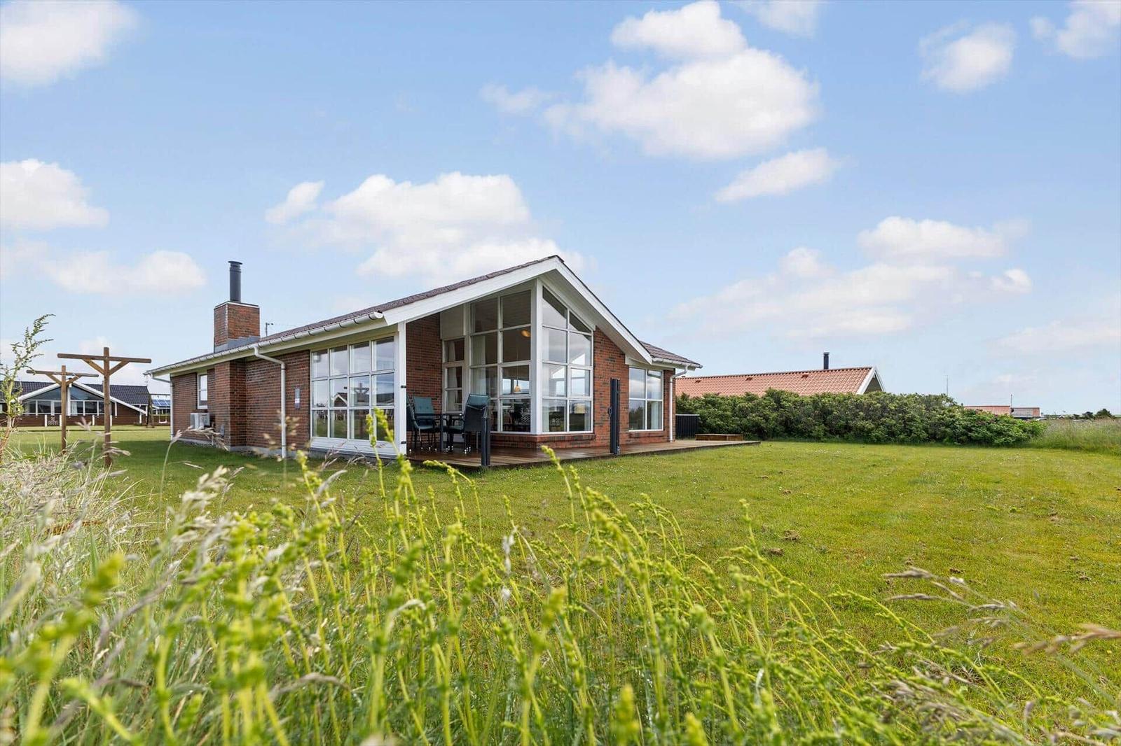 Ein Haus mit großem Glasbereich und Terrasse steht auf einer grünen Wiese unter blauem Himmel.