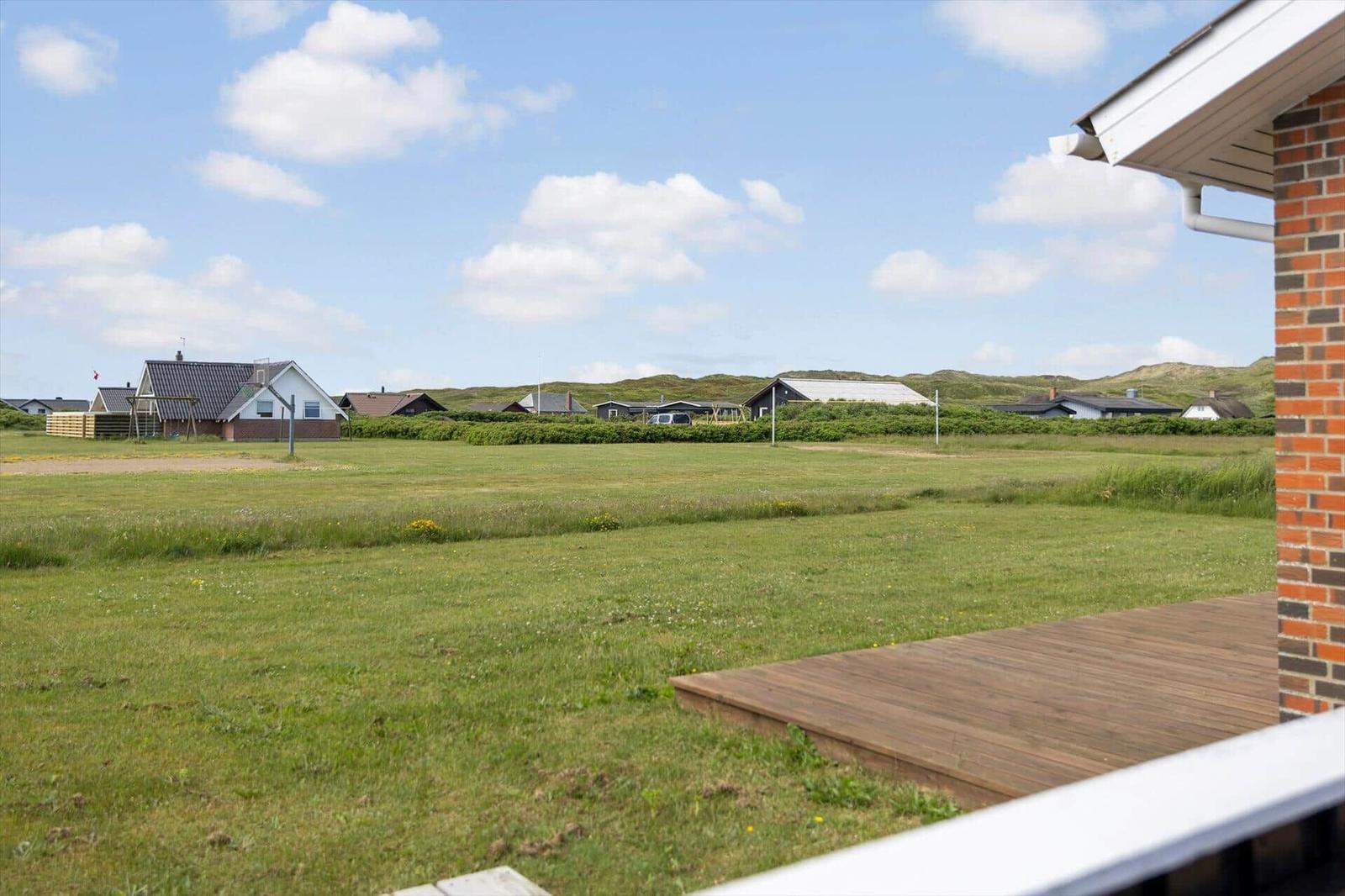 Grünes Feld mit Blick auf Häuser und Dünen. Holzterrasse und roter Ziegelstein sichtbar.